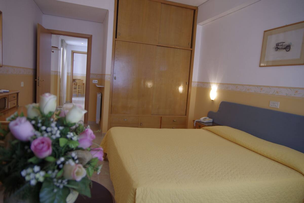 Una habitación de hotel con una cama, un armario de madera, una mesita de noche, un jarrón con flores y un cuadro en la pared.
