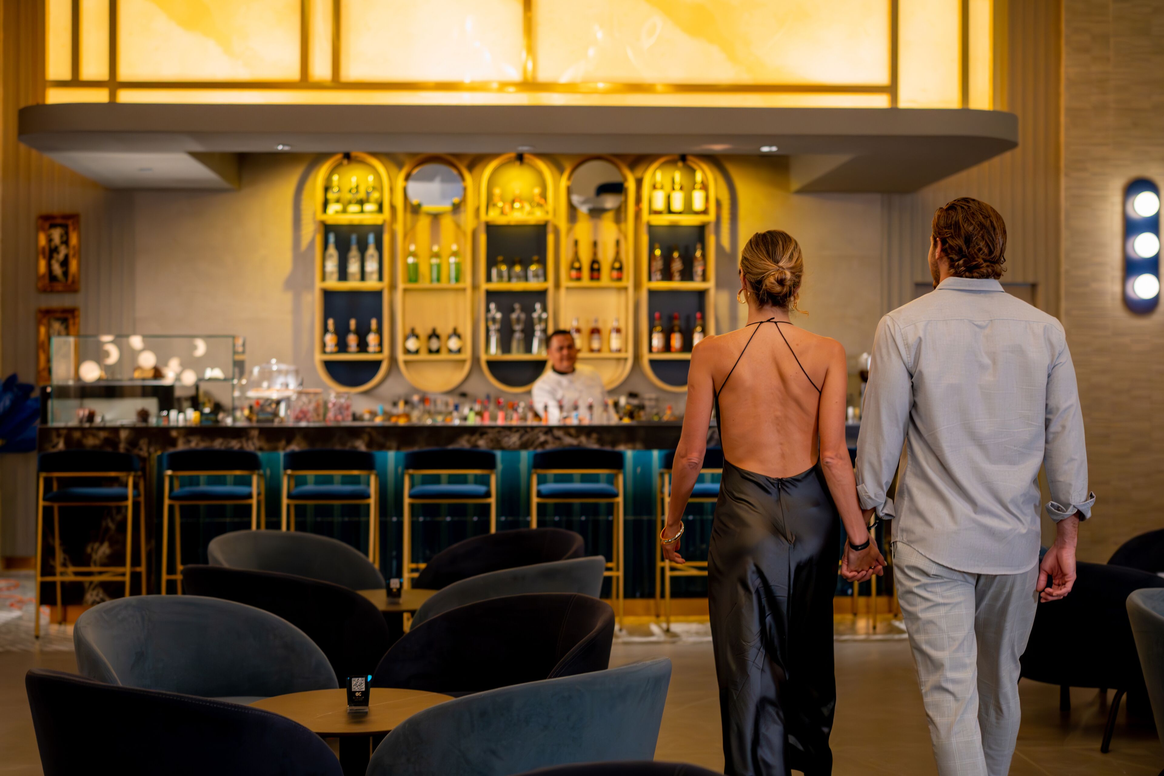 A bar area with multiple shelves of bottles, a bartender, and seating with tables and chairs.