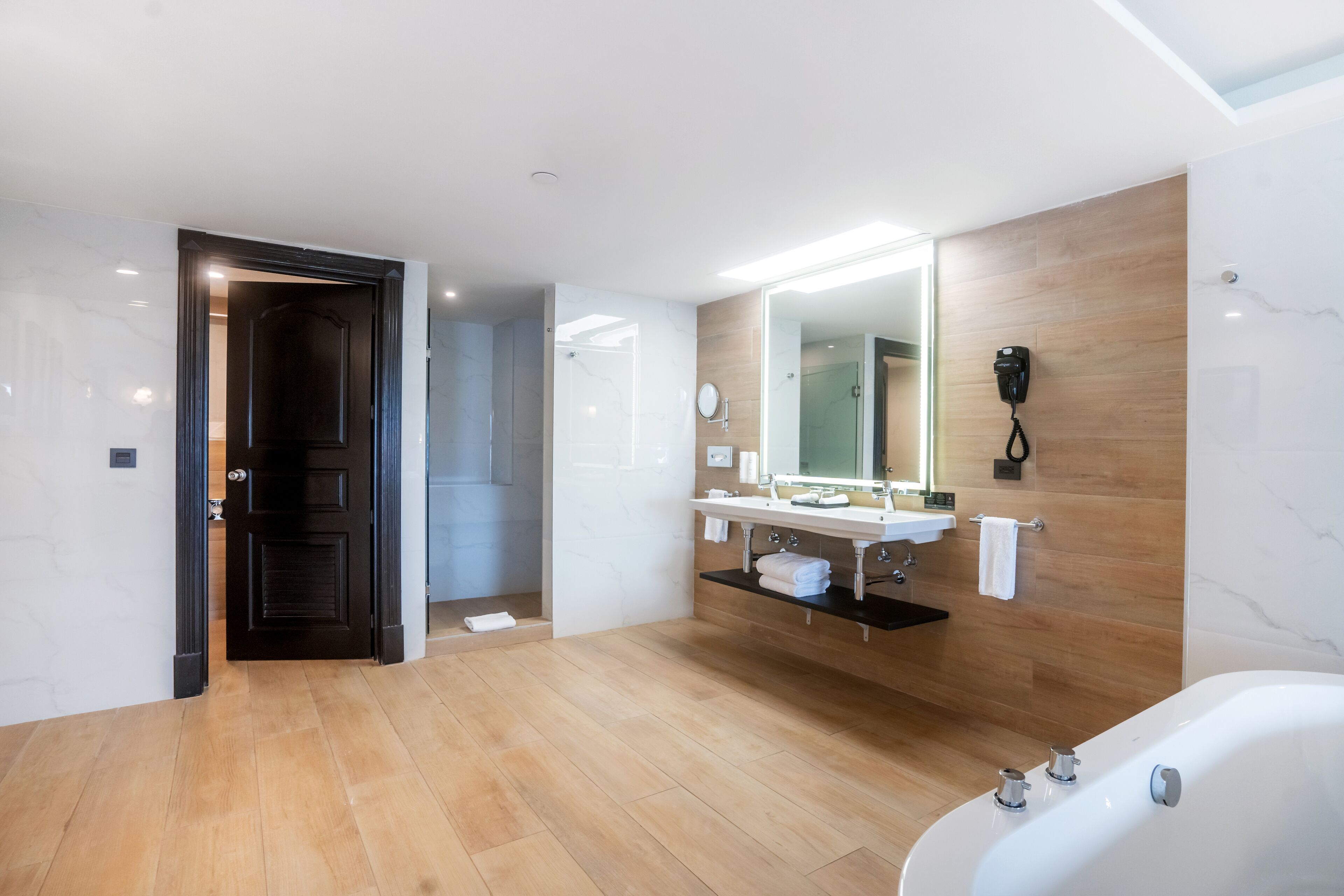 Un baño moderno con suelo de madera, un espejo grande, un lavabo blanco y una bañera.