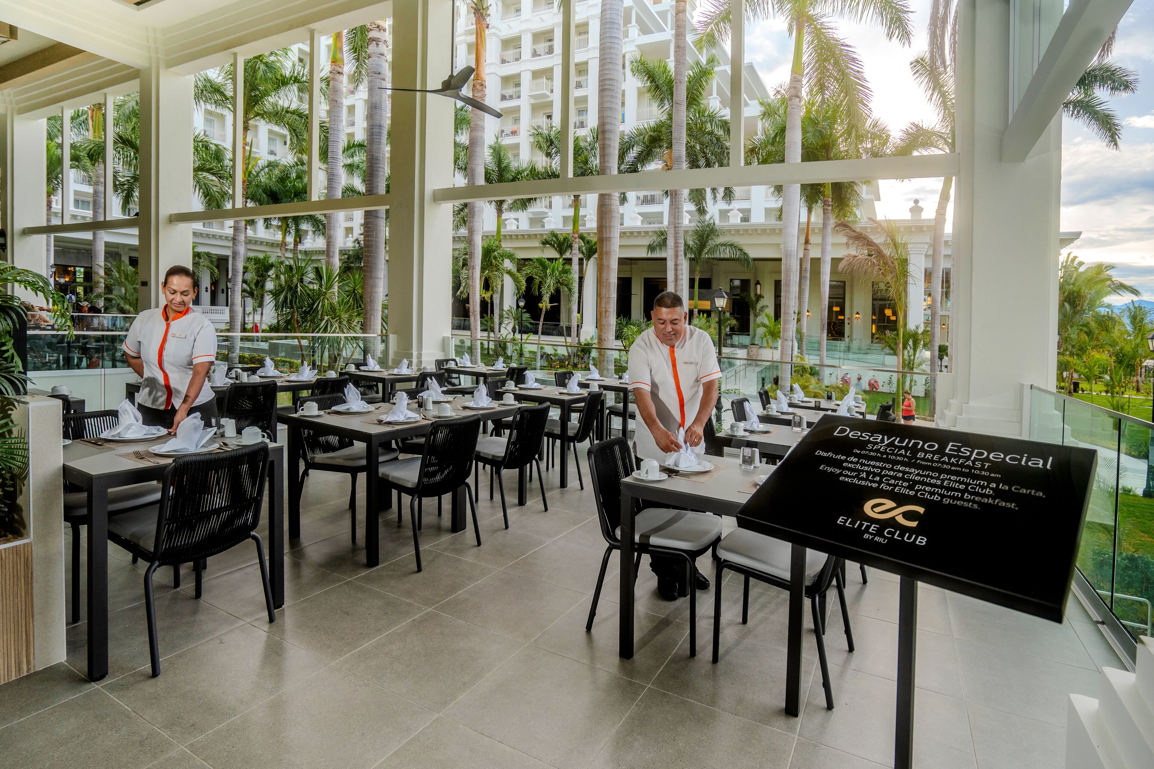 A restaurant with tables set for breakfast, a sign for 'Desayuno Especial', and a view of palm trees and a building in the background.