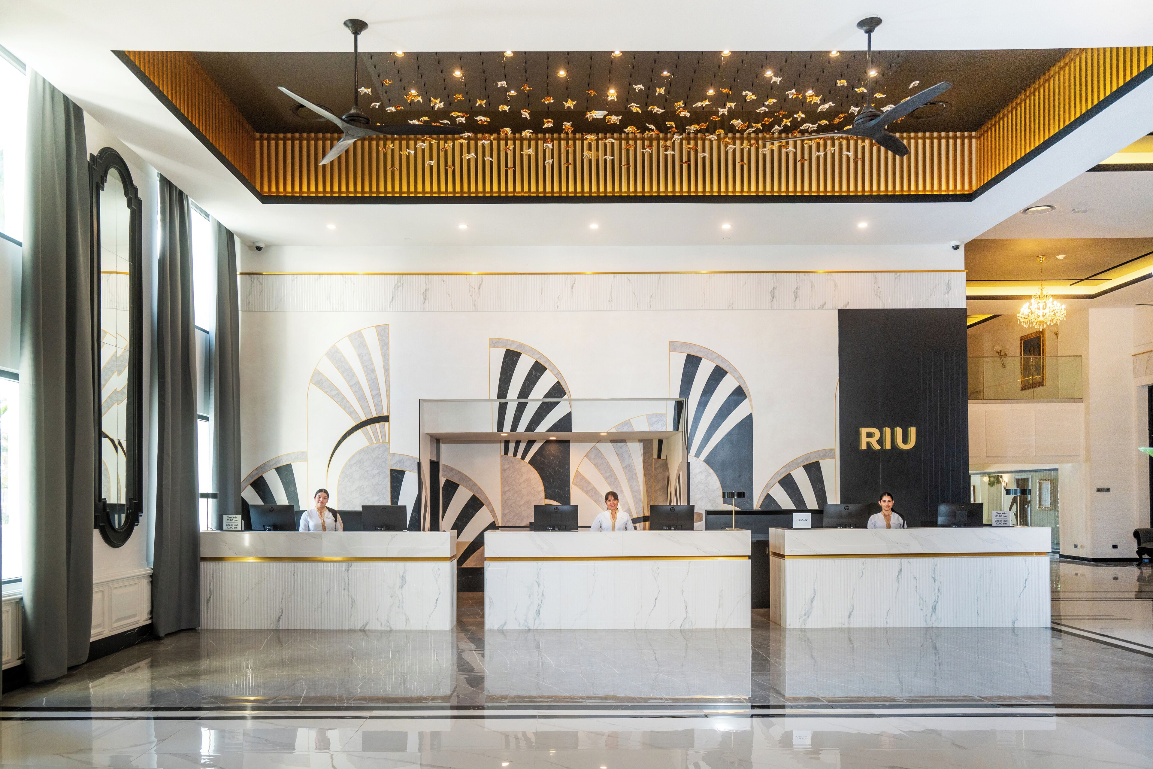 A modern hotel reception area with marble counters, a large RIU sign, and a ceiling with hanging lights.