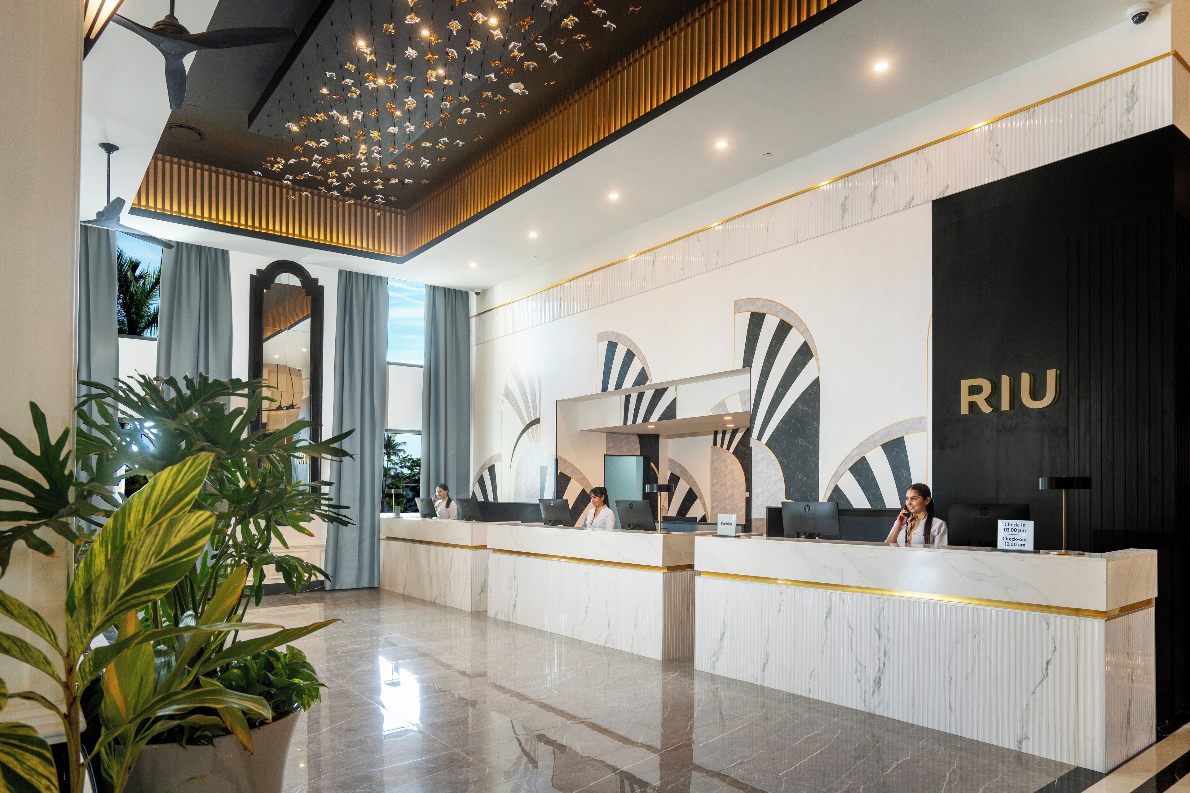 A hotel reception area with a modern design, featuring a marble reception desk, a large black wall with the hotel name, and a ceiling with hanging lights.