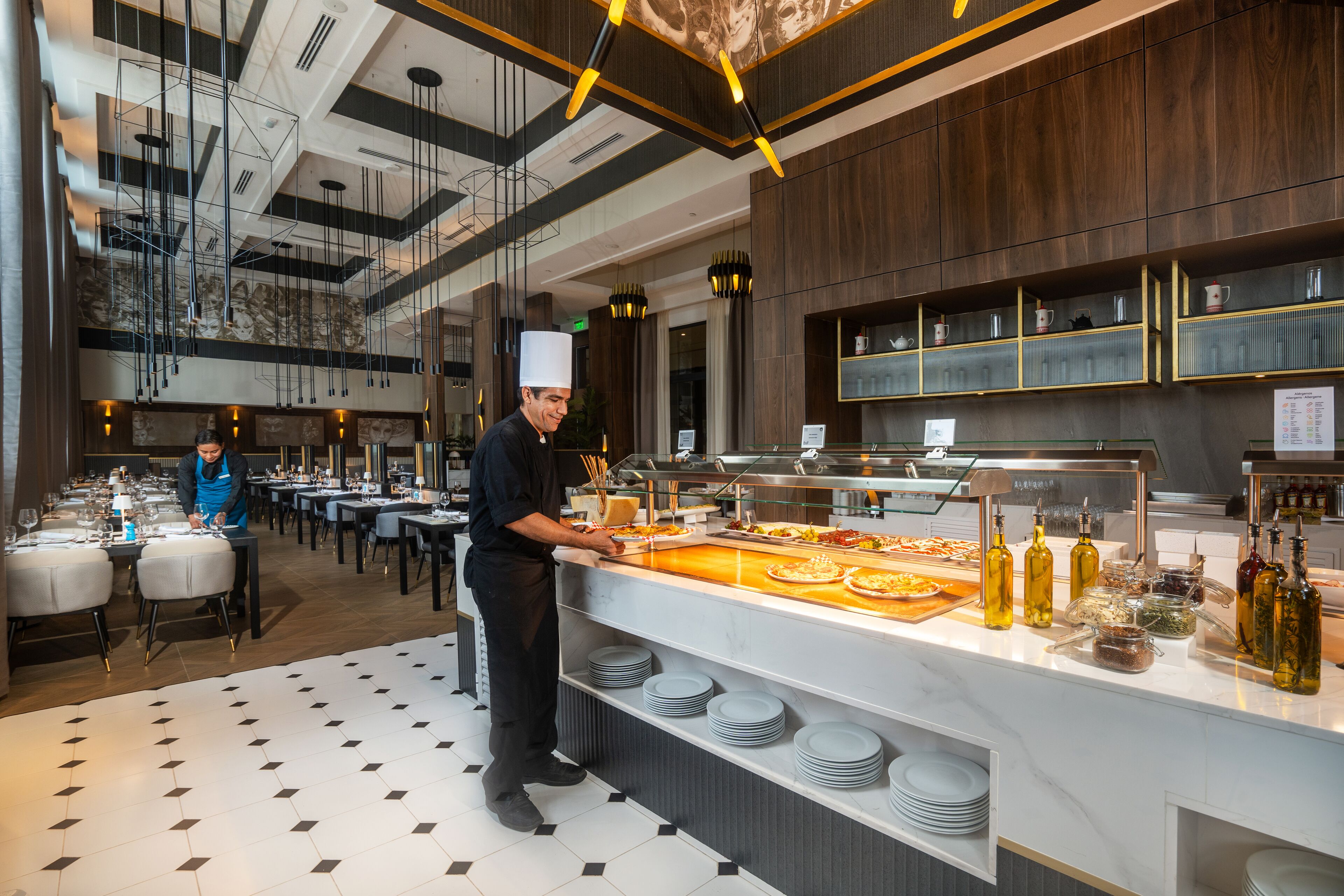 A modern restaurant with a buffet setup, a chef preparing food, and a dining area with tables and chairs.
