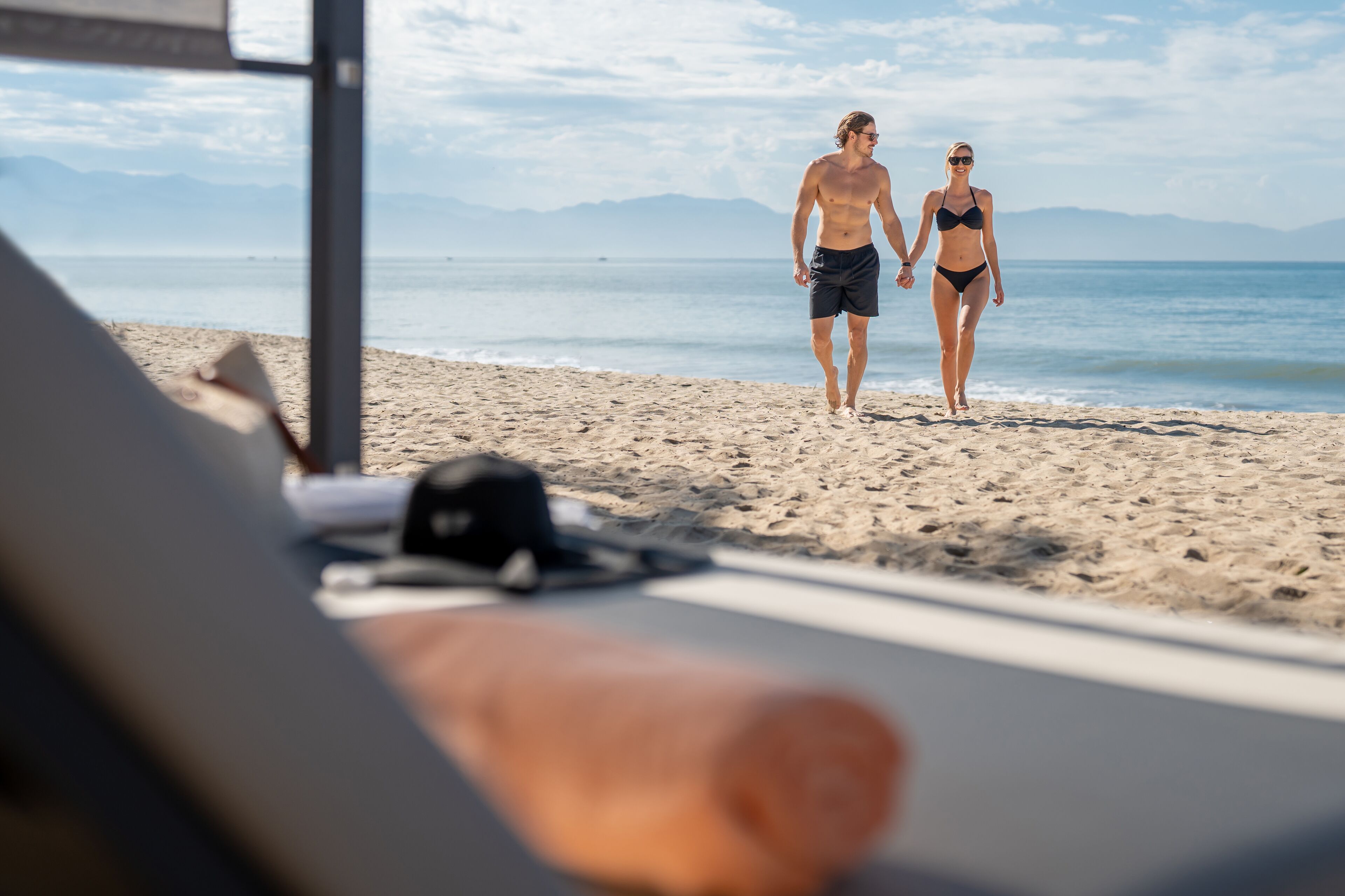 A beach scene with a couple walking on the sand, a beach chair, and a surfboard.
