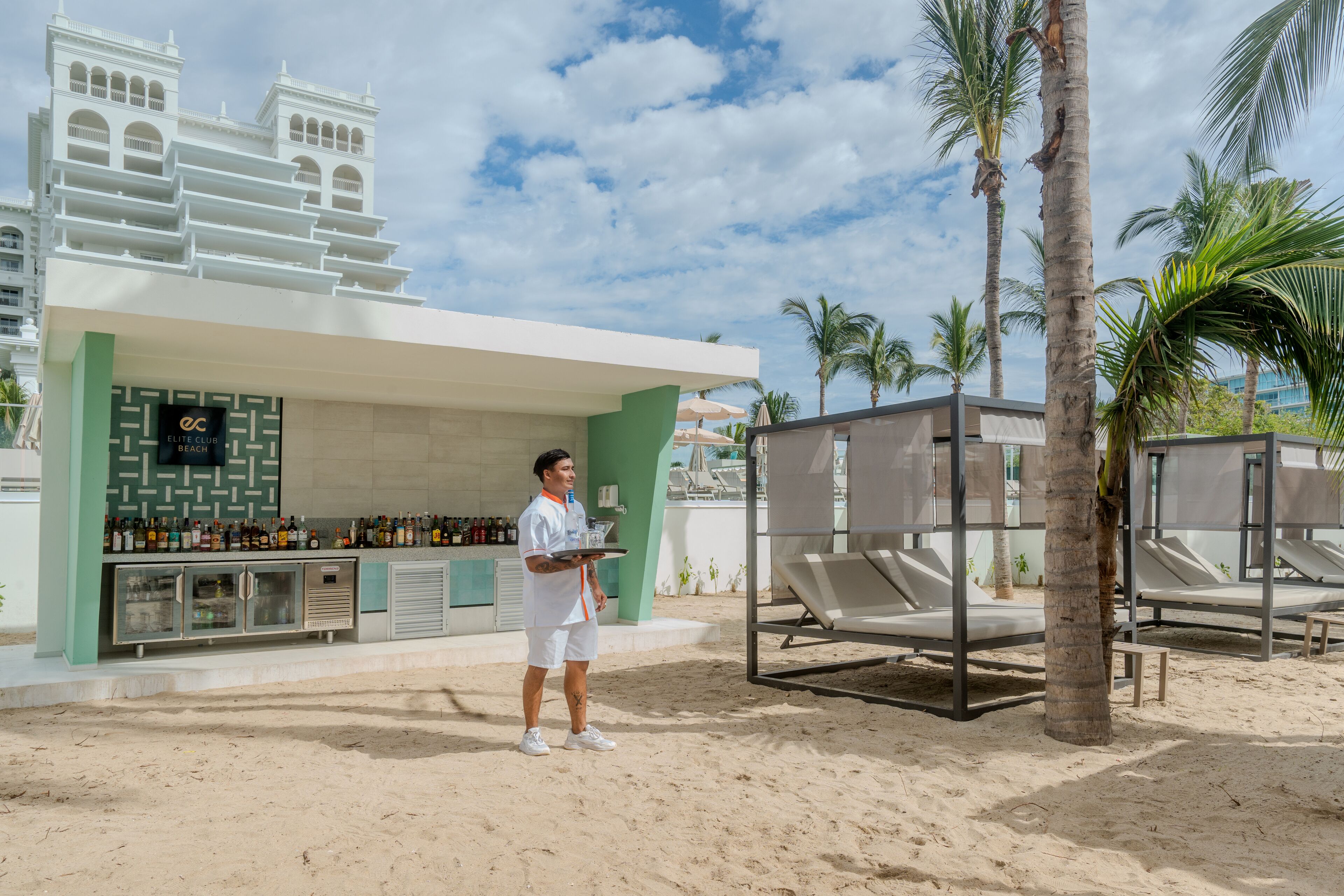A bar area with a counter displaying bottles, a person holding a tray, and lounge chairs under palm trees.