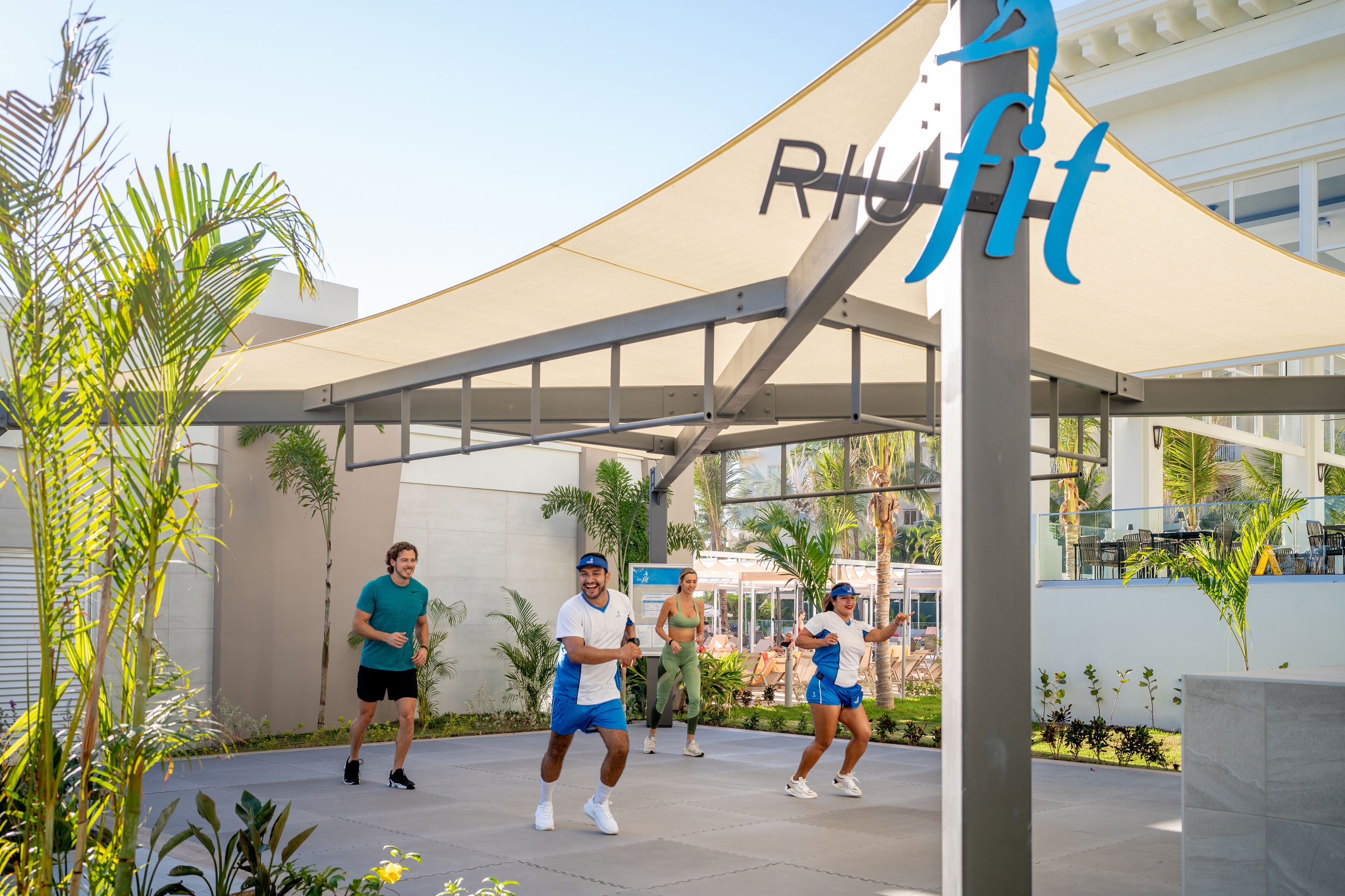 A group of people exercising outdoors in a covered area with a modern architectural design, palm trees, and a clear sky.