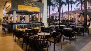Una zona de bar con un letrero 'Baleró', rodeada de sillas y mesas de mimbre, con vista a palmeras y un edificio al fondo.