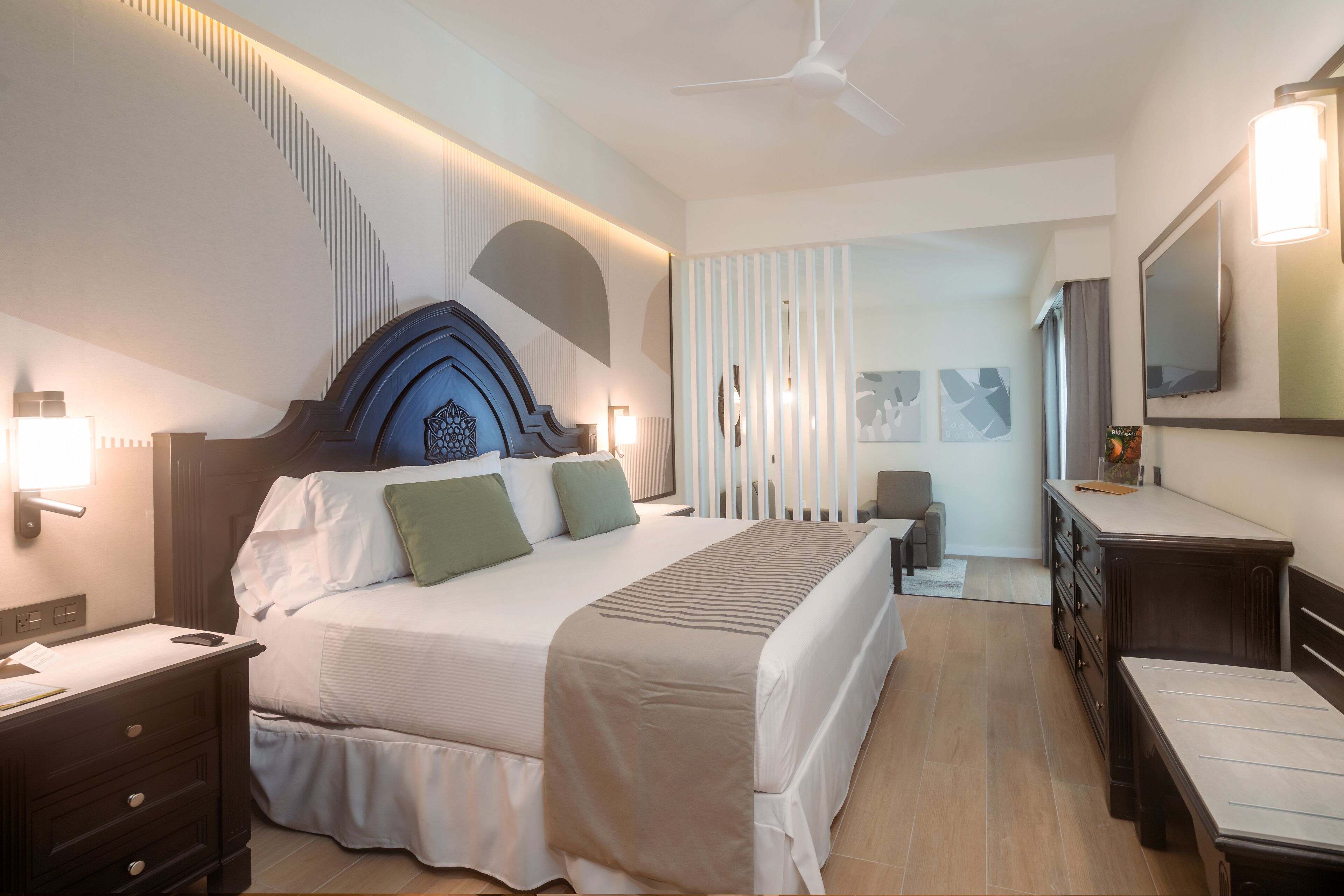 A modern hotel room with a large bed, bedside tables, a dresser, a desk, and a ceiling fan.