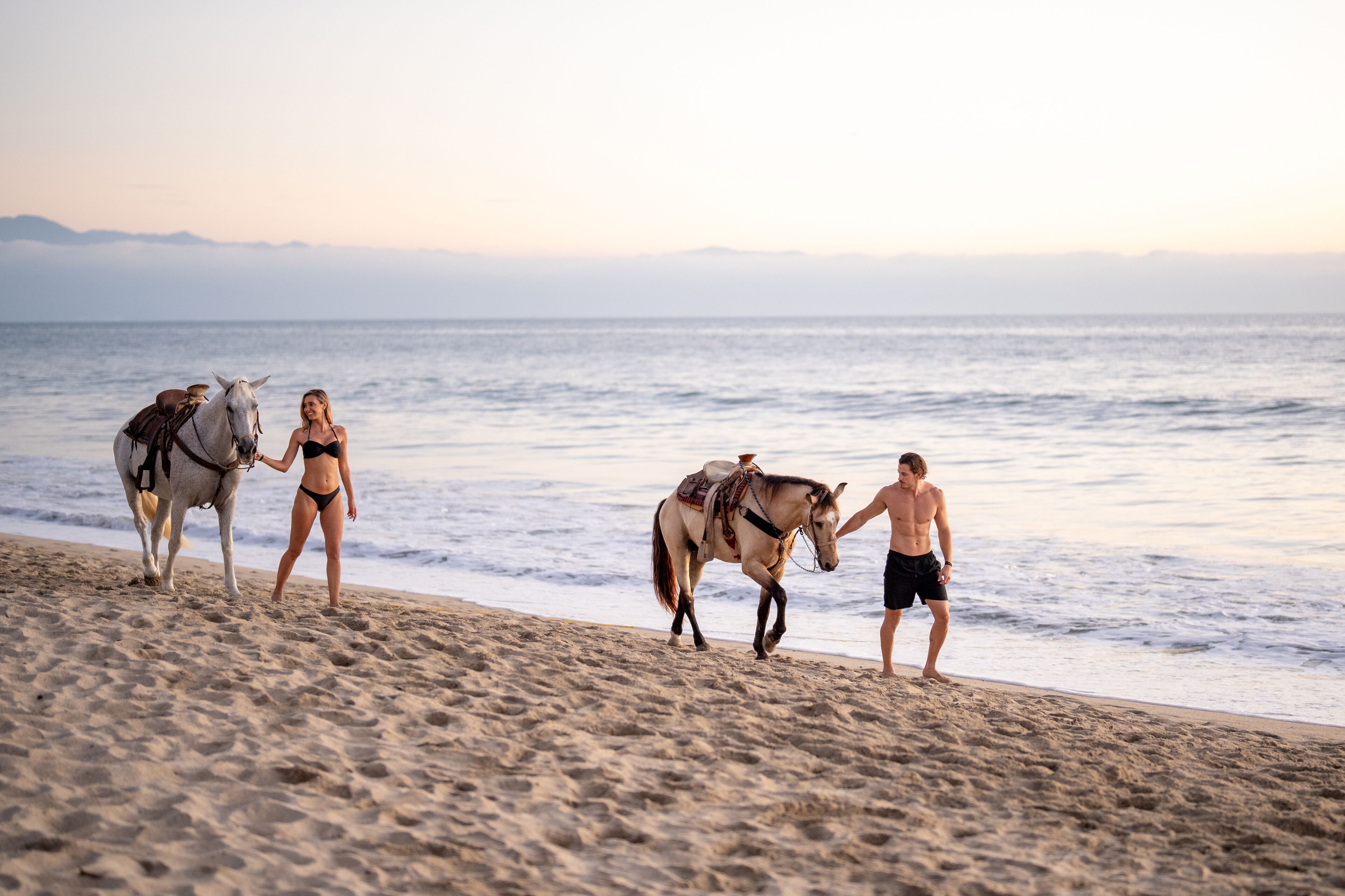 A beach scene with two horses and two people.