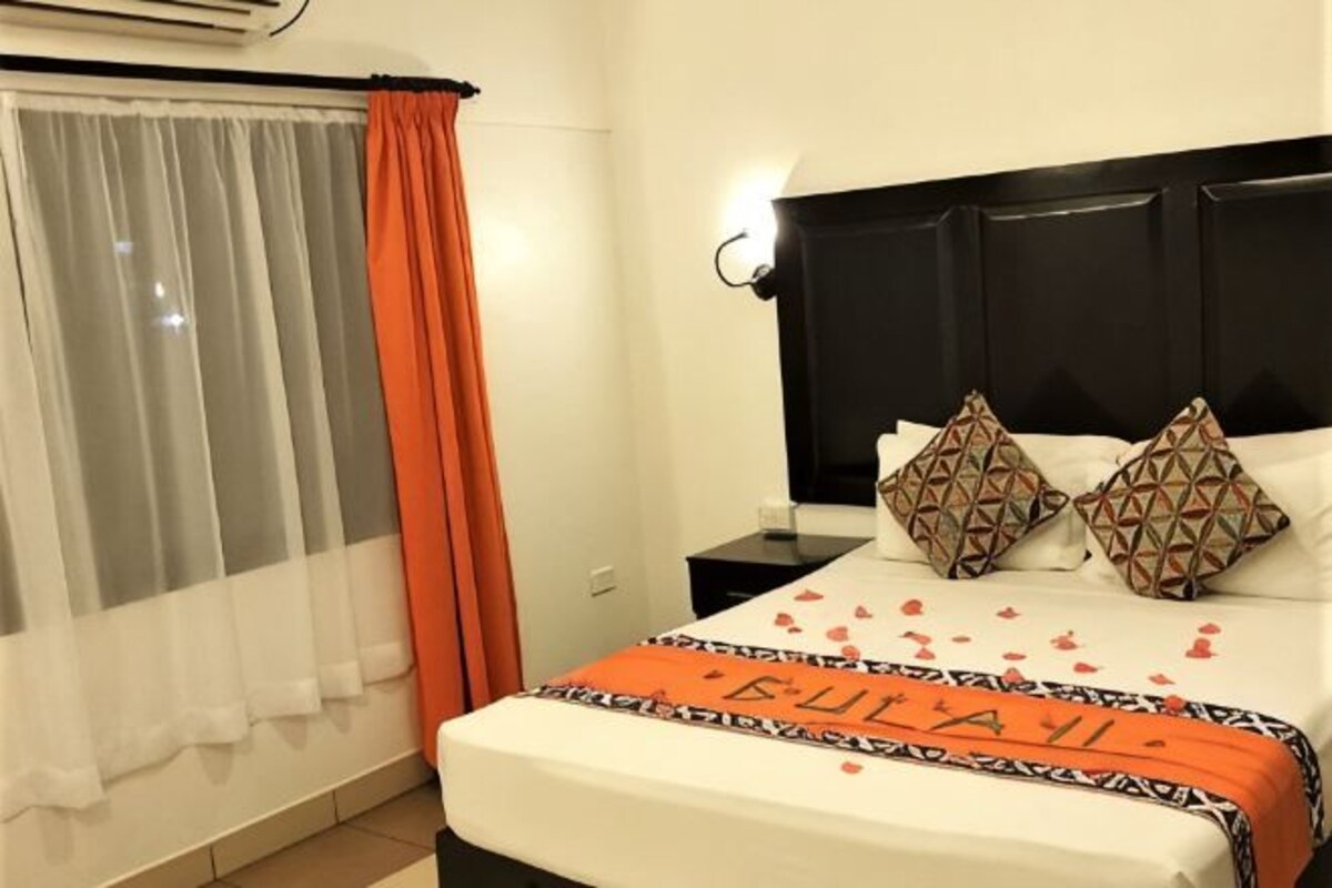 A hotel room with a bed, a nightstand, a wall-mounted lamp, a window with curtains, and a bedspread with a name.