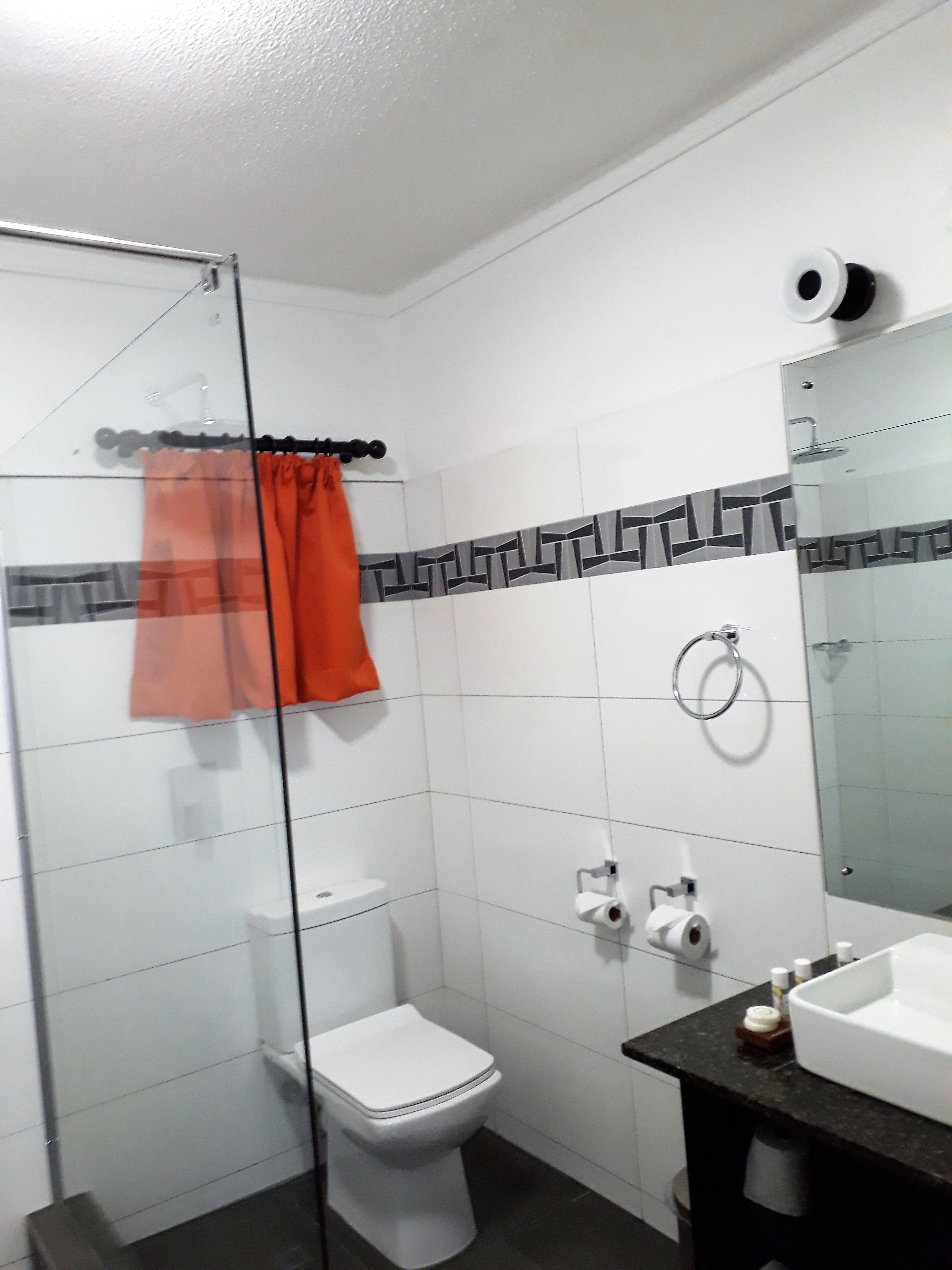 Un baño con inodoro, mampara de ducha de vidrio, lavamanos con espejo y toallero.