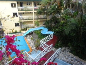 A pool area with a blue slide, surrounded by lush greenery and a multi-story building with balconies.
