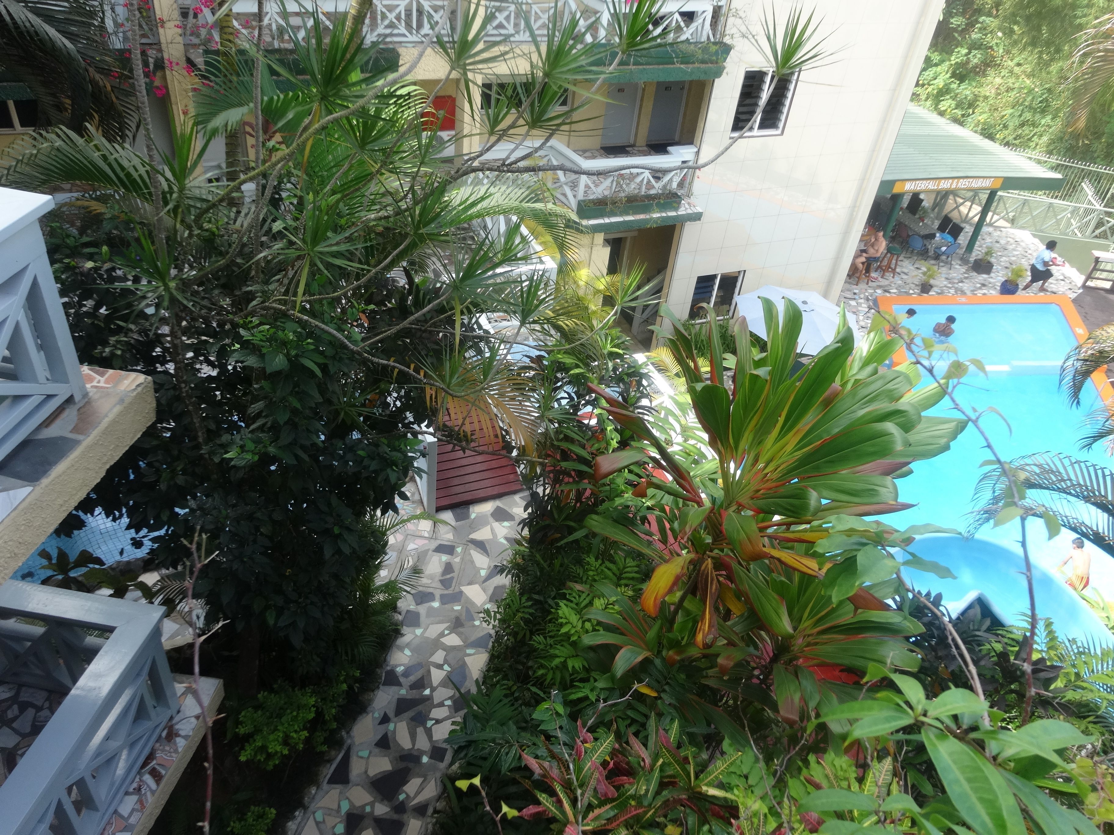 A pool area with a tiled deck, surrounded by lush greenery and a building with a balcony.