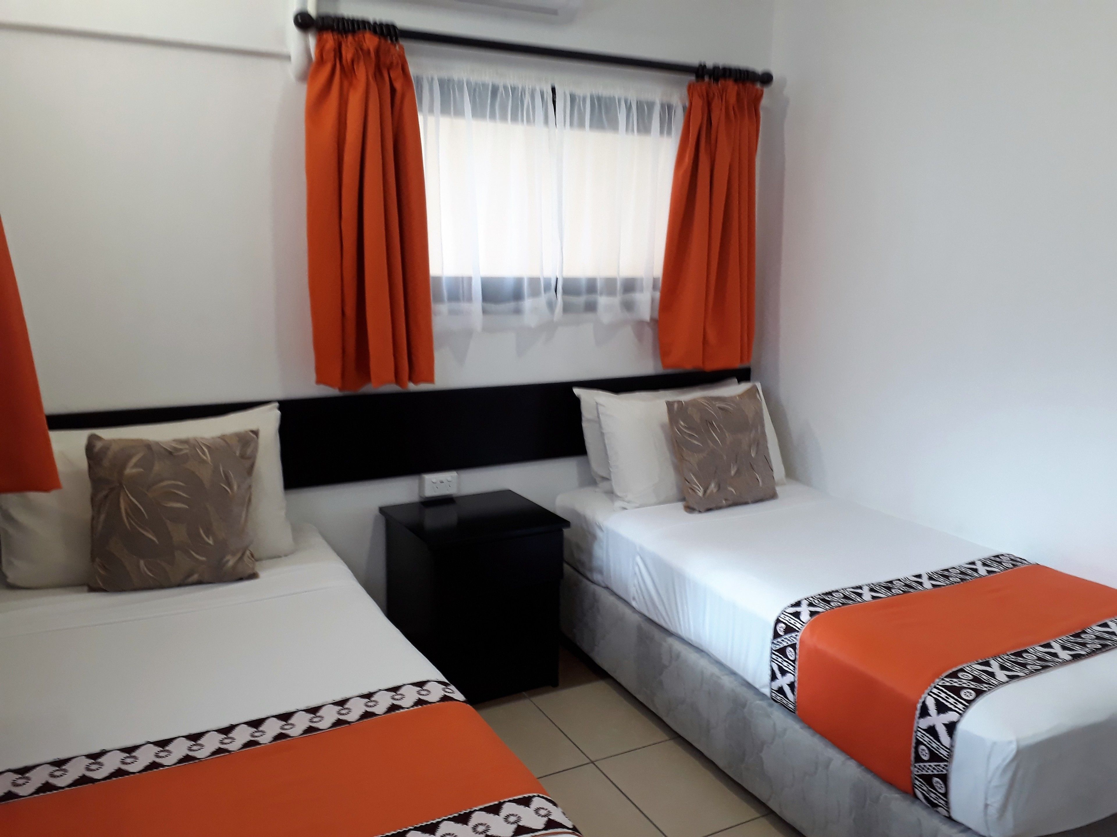 Habitación de hotel con dos camas individuales, una mesita de noche negra y una ventana con cortinas naranjas.