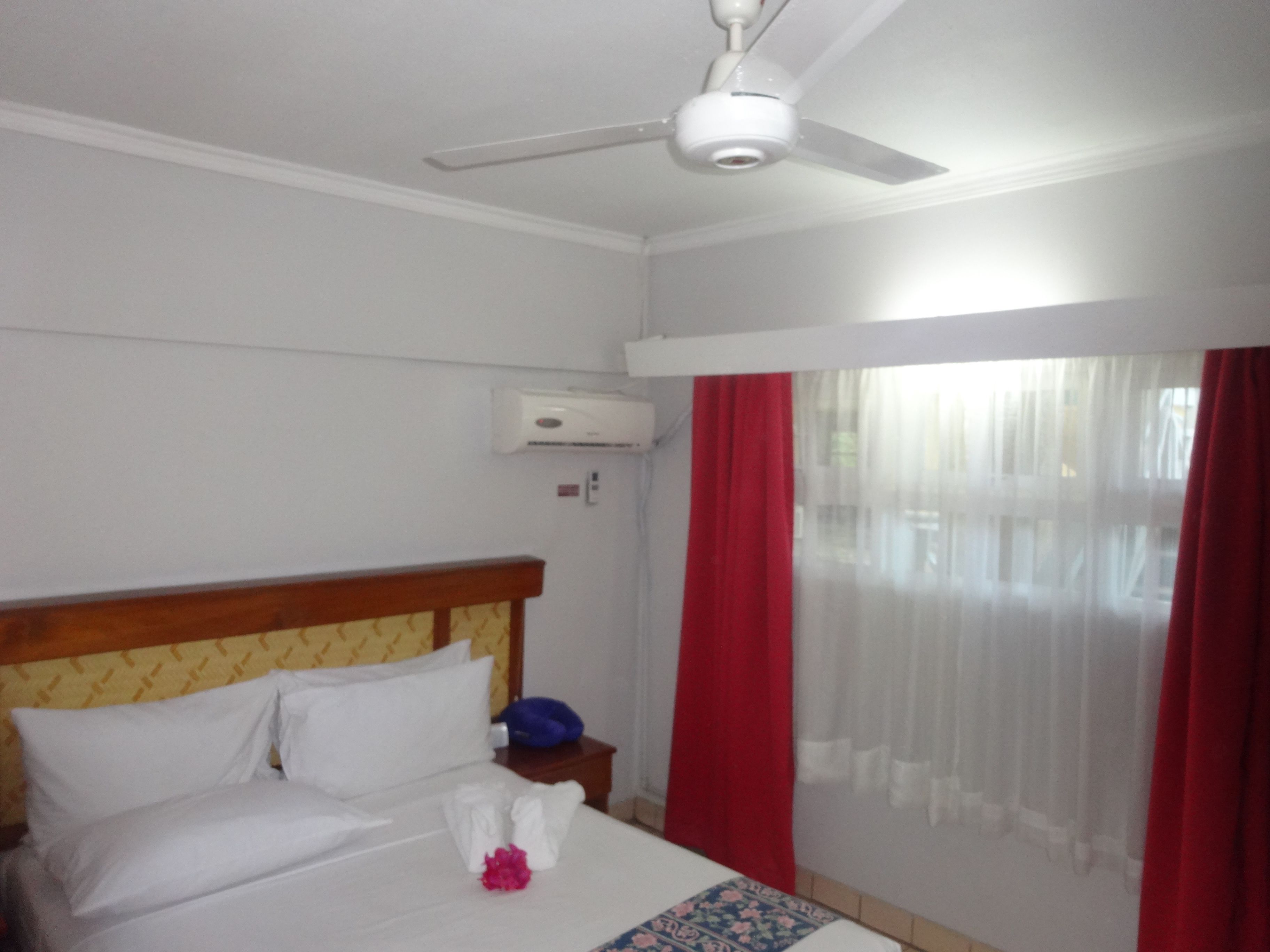 A hotel room with a bed, a ceiling fan, an air conditioning unit, a nightstand, and a window with red curtains.