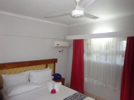 Habitación de hotel con cama, ventilador de techo, equipo de aire acondicionado, mesita de noche y ventana con cortinas rojas.