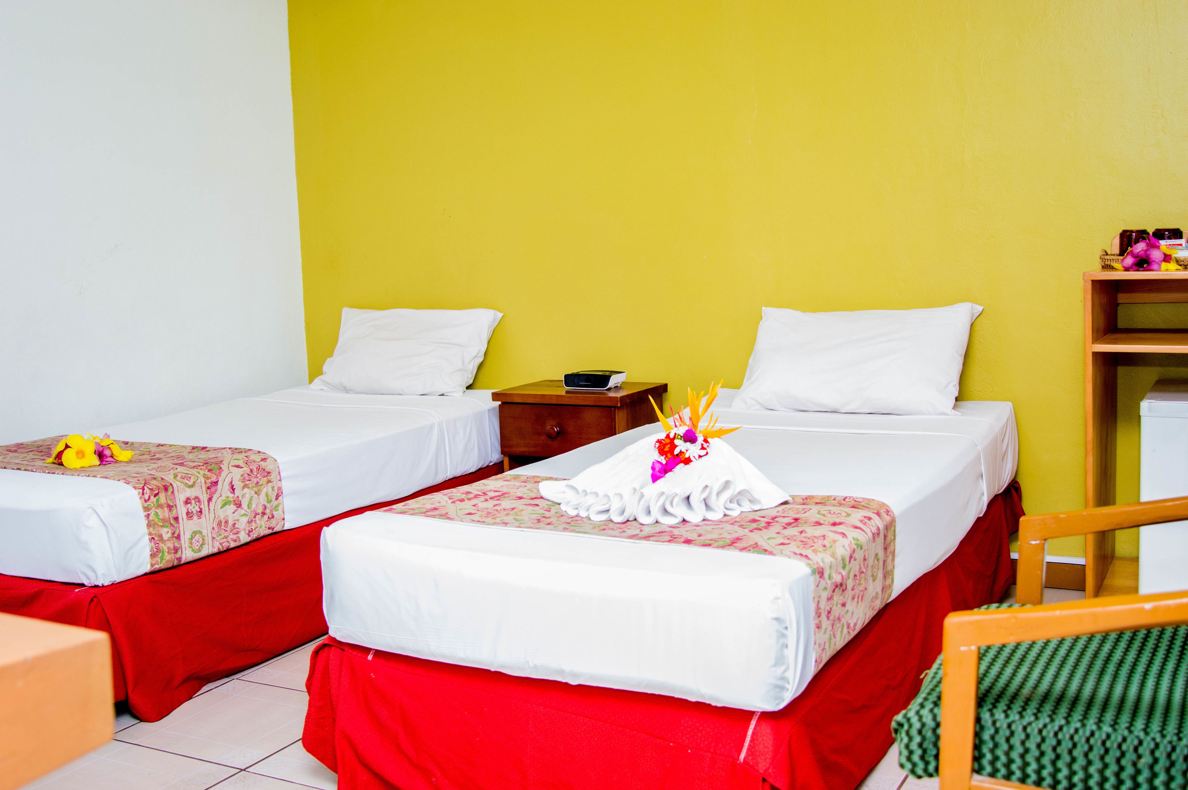 Habitación de hotel con dos camas, cada una con falda roja y ropa blanca. Hay una mesita de noche de madera entre las camas con un objeto negro pequeño encima.