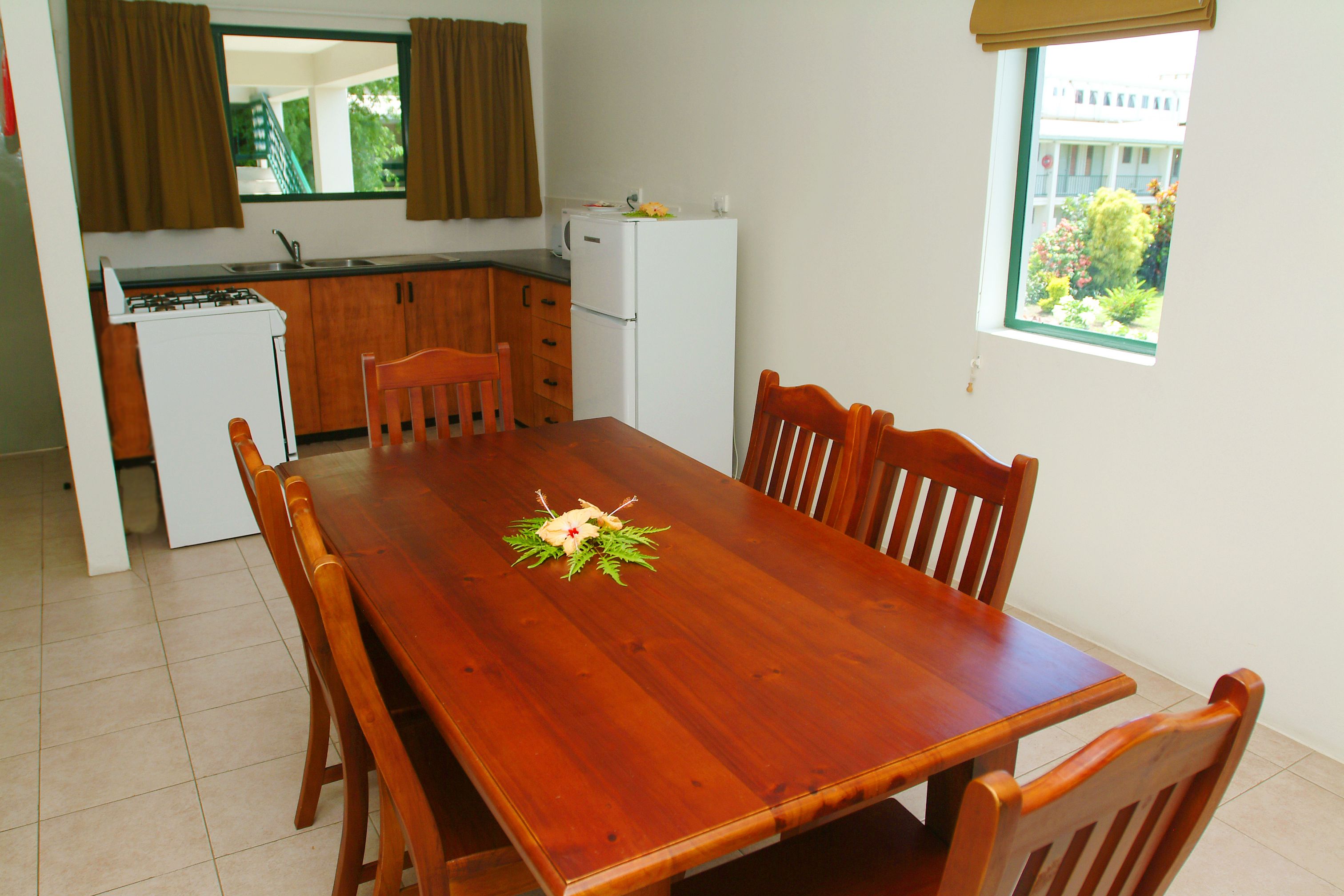 A dining area with a wooden table and chairs, a small kitchen with a refrigerator and stove, and a window with a view of greenery.
