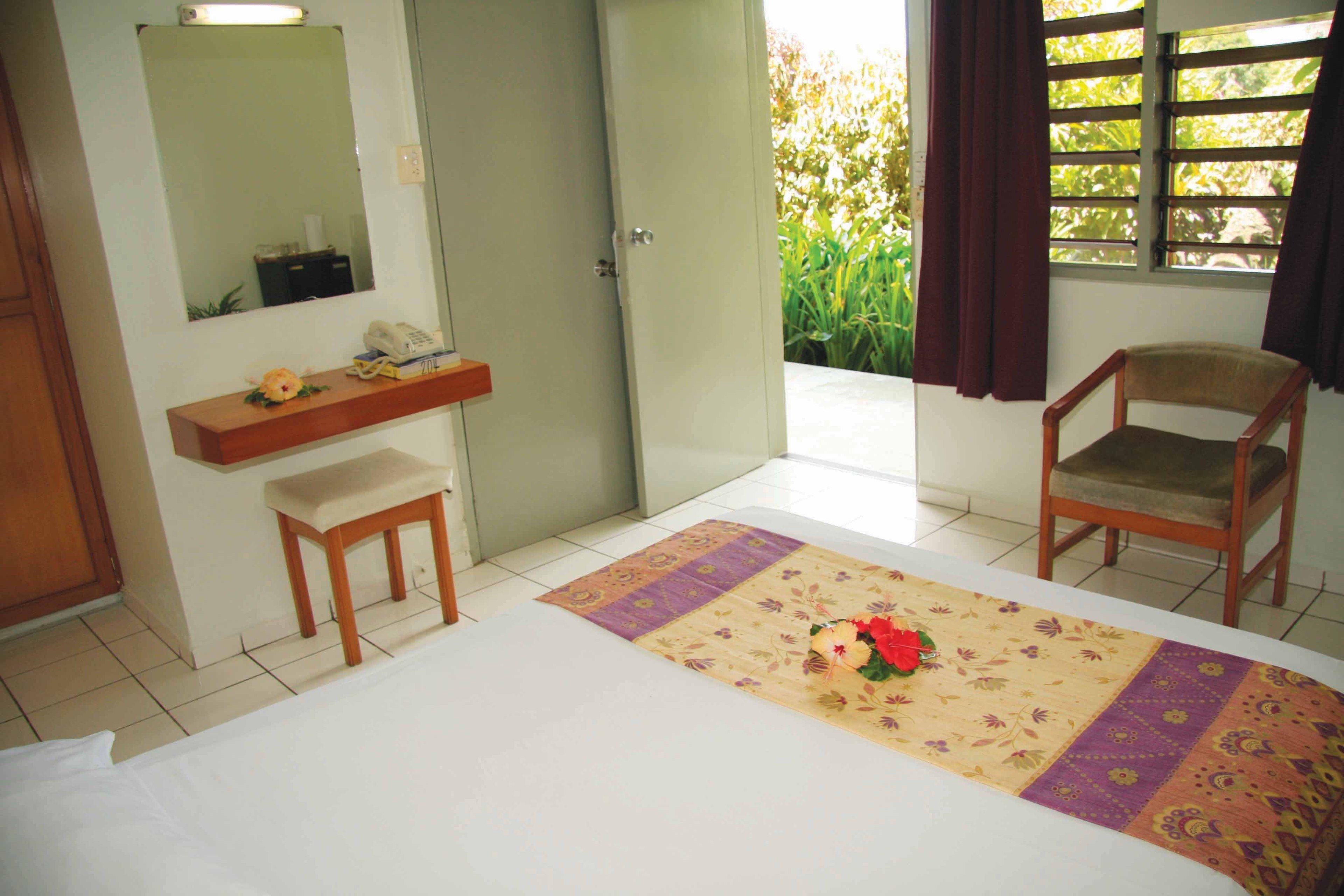 Quarto com cama, cadeira de madeira, mesinha, espelho e janela com cortinas.