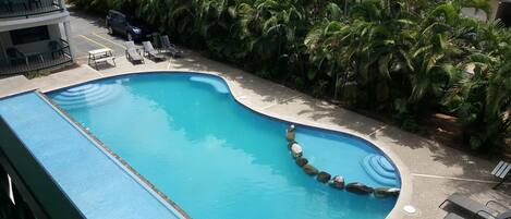 A swimming pool with a curved edge, surrounded by palm trees and a building with balconies.