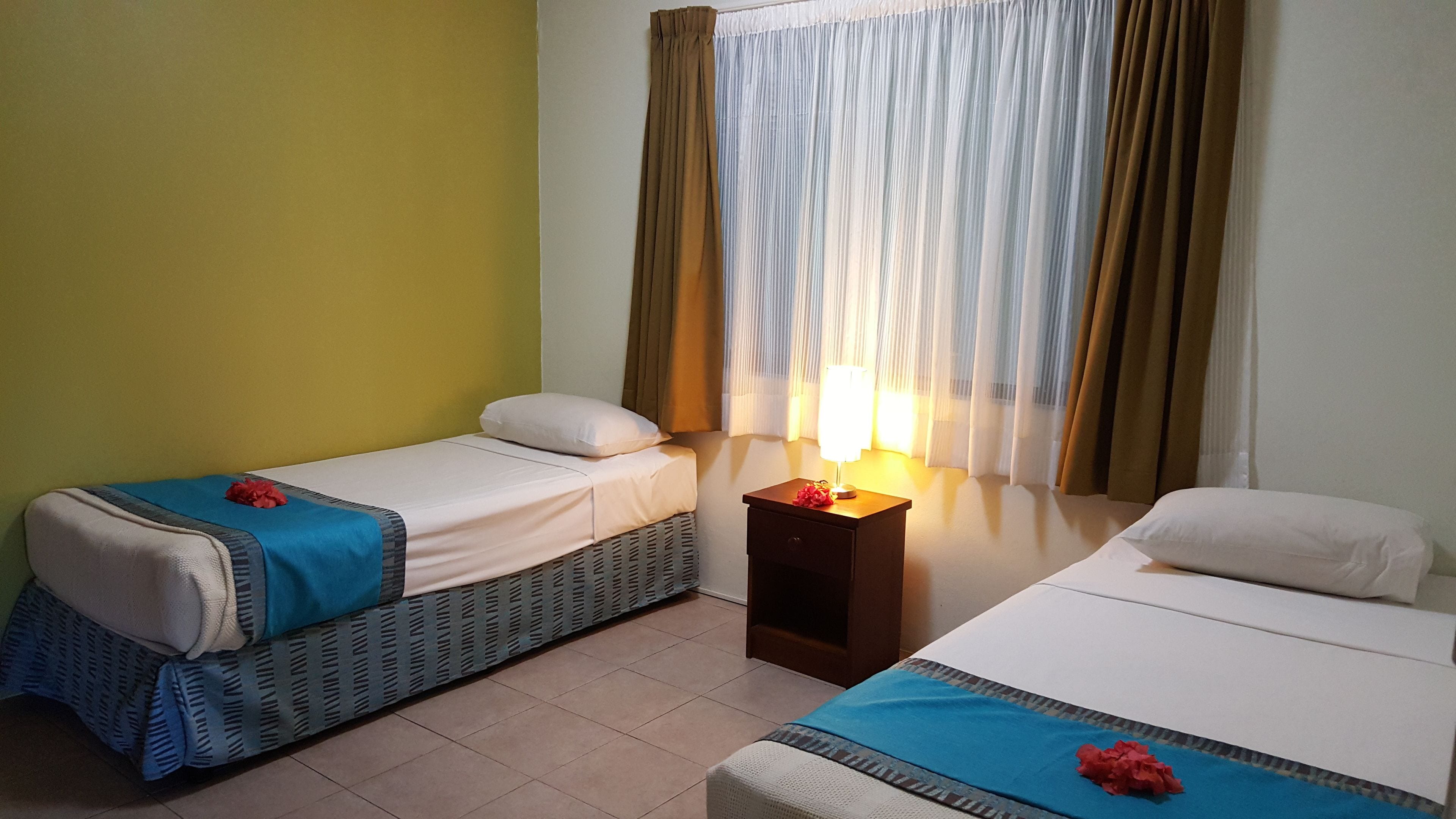 A hotel room with two single beds, a nightstand with a lamp, and a window with curtains.