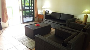 A living room with a black sofa, wicker furniture, a coffee table with a red flower, and a view through a glass door. - Grand West's Villas (Nadi)