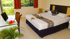 A hotel room with a bed, a desk with a laptop, a chair, a window with curtains, and a view of greenery outside. - Grand West's Villas (Nadi)