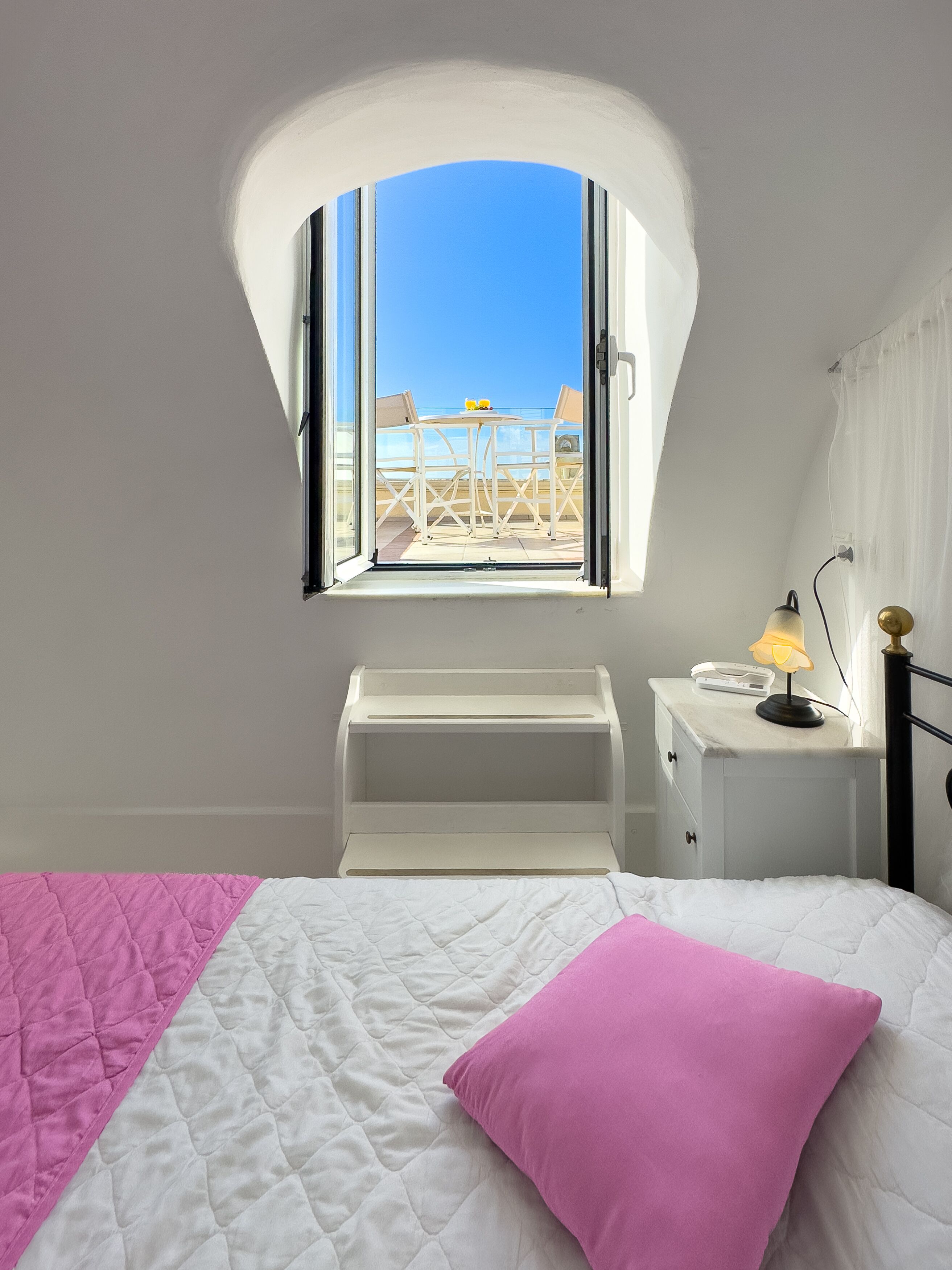 A bedroom with a bed, a window, a bedside table, and a view of an outdoor seating area.
