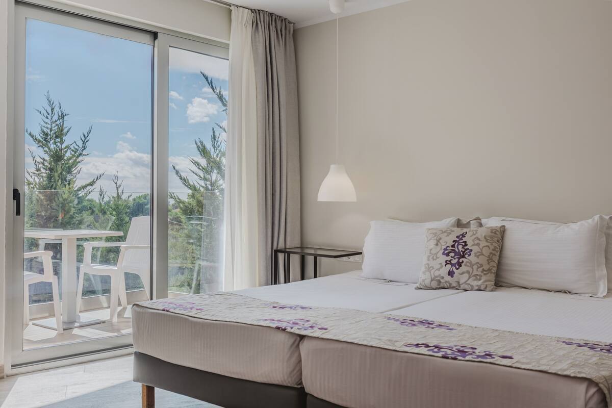 Une chambre avec un grand lit, une table de chevet, une lampe et une vue sur les arbres, visible à travers une porte-fenêtre coulissante.