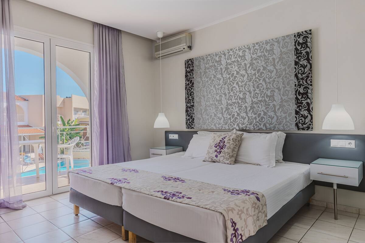 Une chambre d’hôtel avec un lit, deux tables de chevet, une petite table, une grande fenêtre avec des rideaux et une vue sur une piscine.
