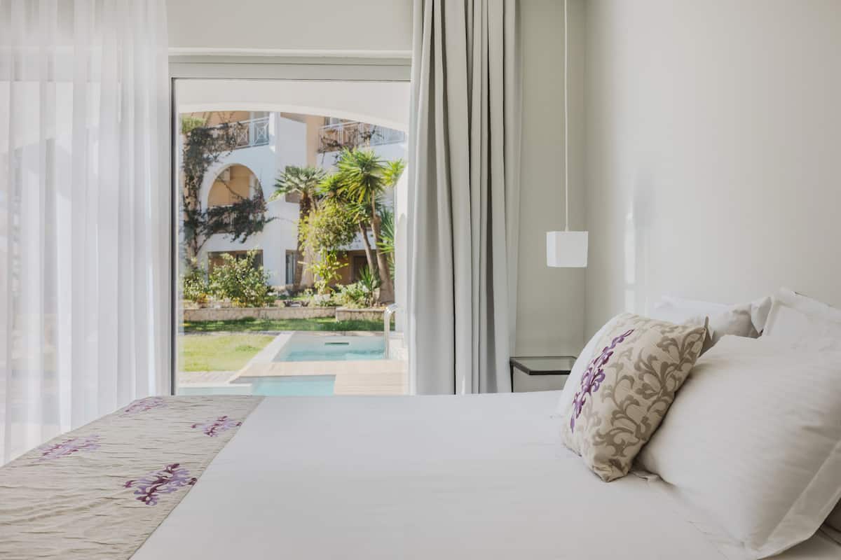 Une chambre d’hôtel avec un lit, une vue sur une piscine extérieure et une cour intérieure agrémentée de plantes et d’éléments architecturaux.