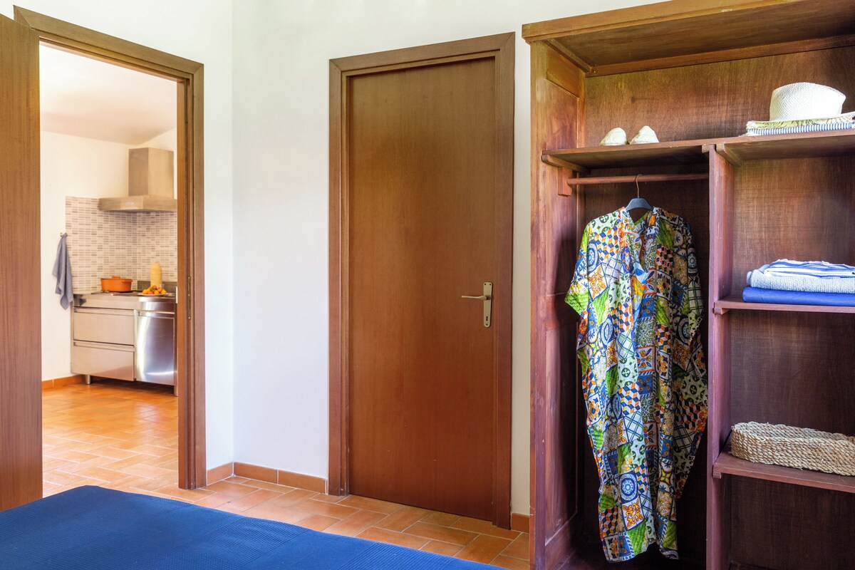 Habitació amb una porta de fusta, un estri de color blau, un teixit colorit penjant i una cuina visible a través d'una porta oberta.