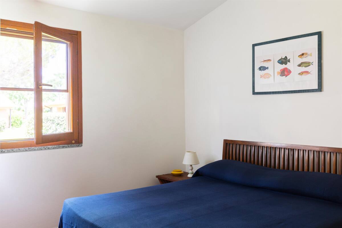 Une chambre avec un lit, une table de chevet avec une lampe, une illustration encadrée représentant un poisson accrochée au mur et une fenêtre avec des stores.