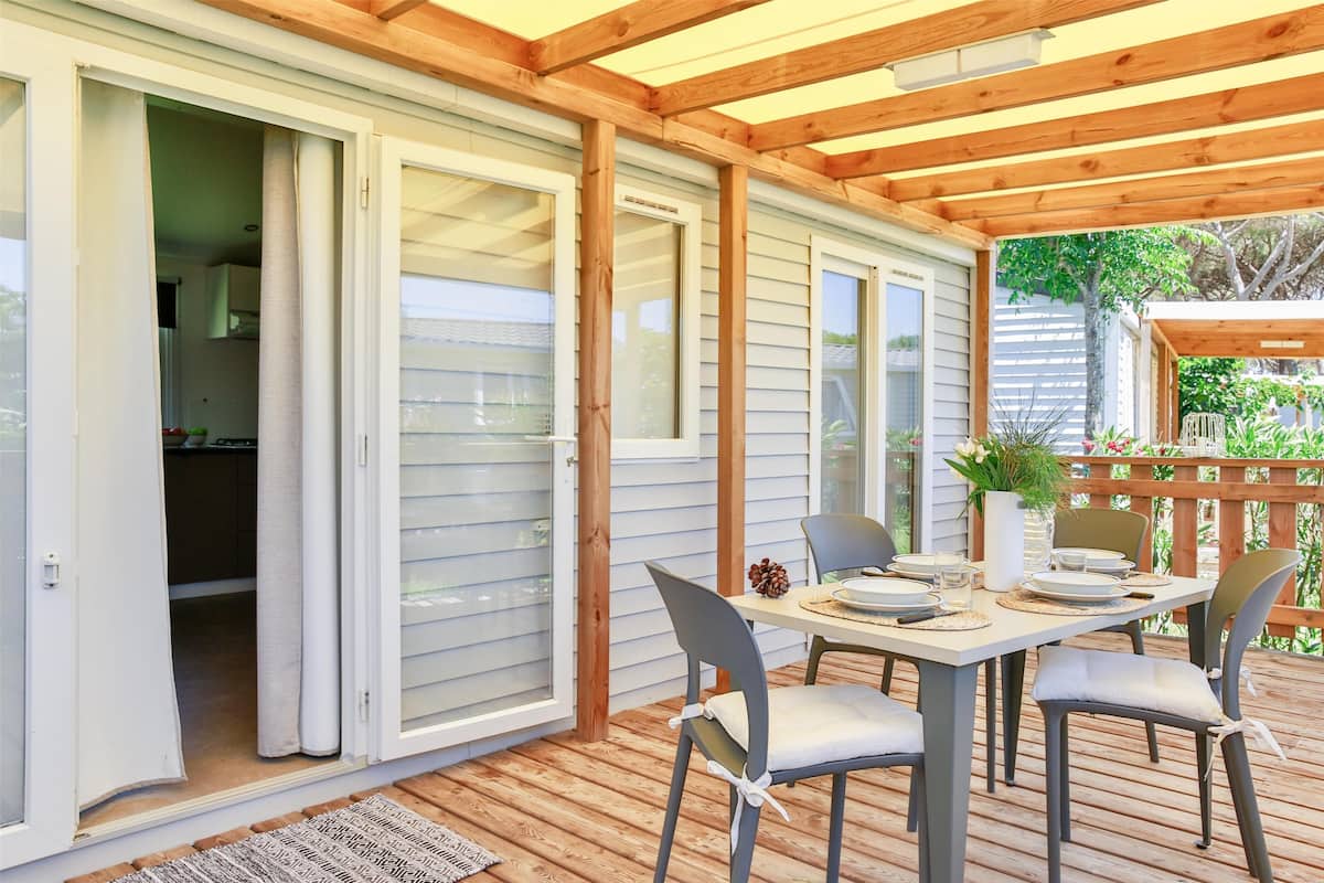 Une terrasse couverte avec une table dressée pour deux personnes, des portes-fenêtres coulissantes donnant sur une cuisine et une terrasse en bois dotée d’une rambarde.