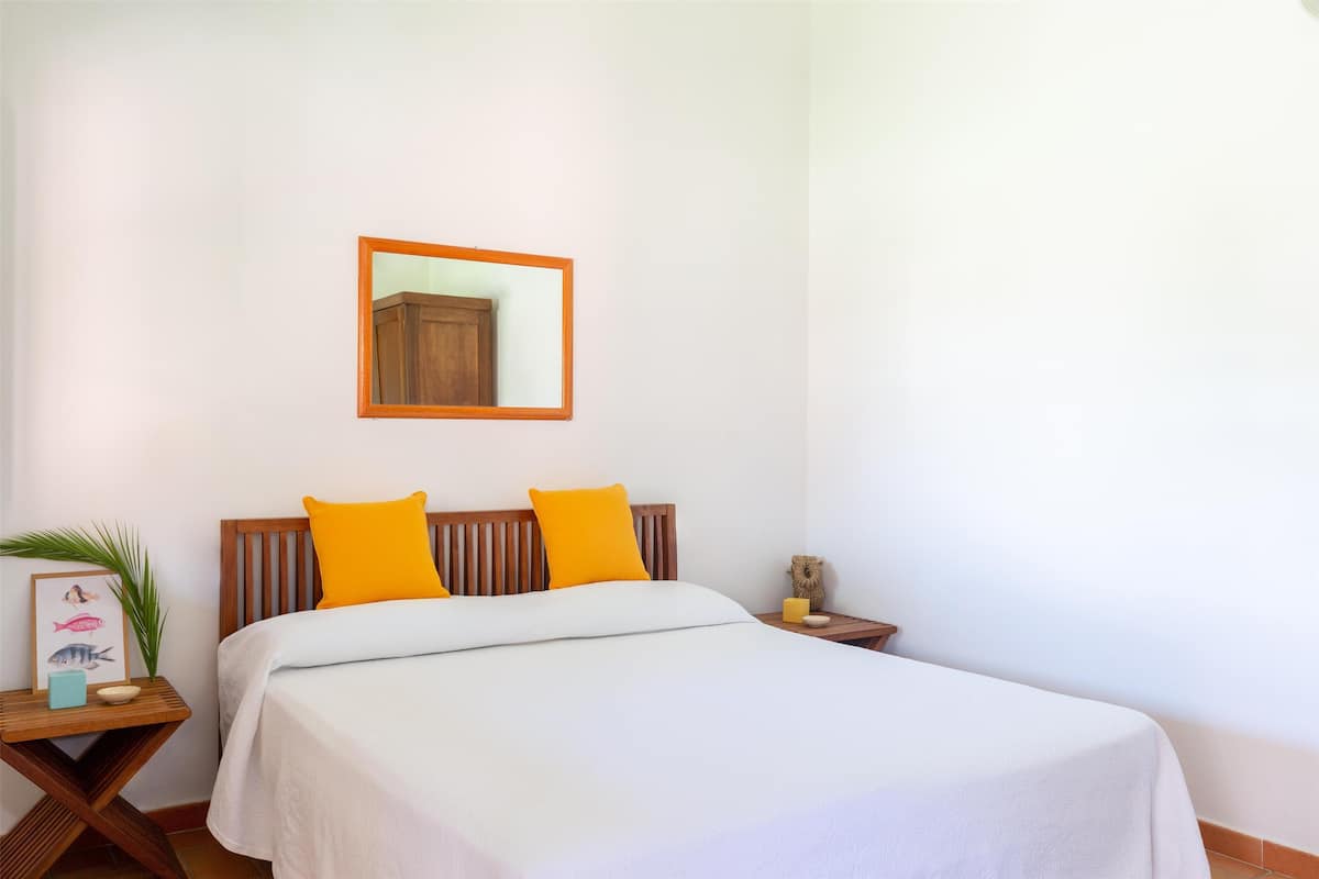 Un lit bien fait, avec une literie blanche et deux coussins jaune vif, une tête de lit en bois, une petite table de chevet avec une lampe et, au mur, un tableau représentant des poissons.