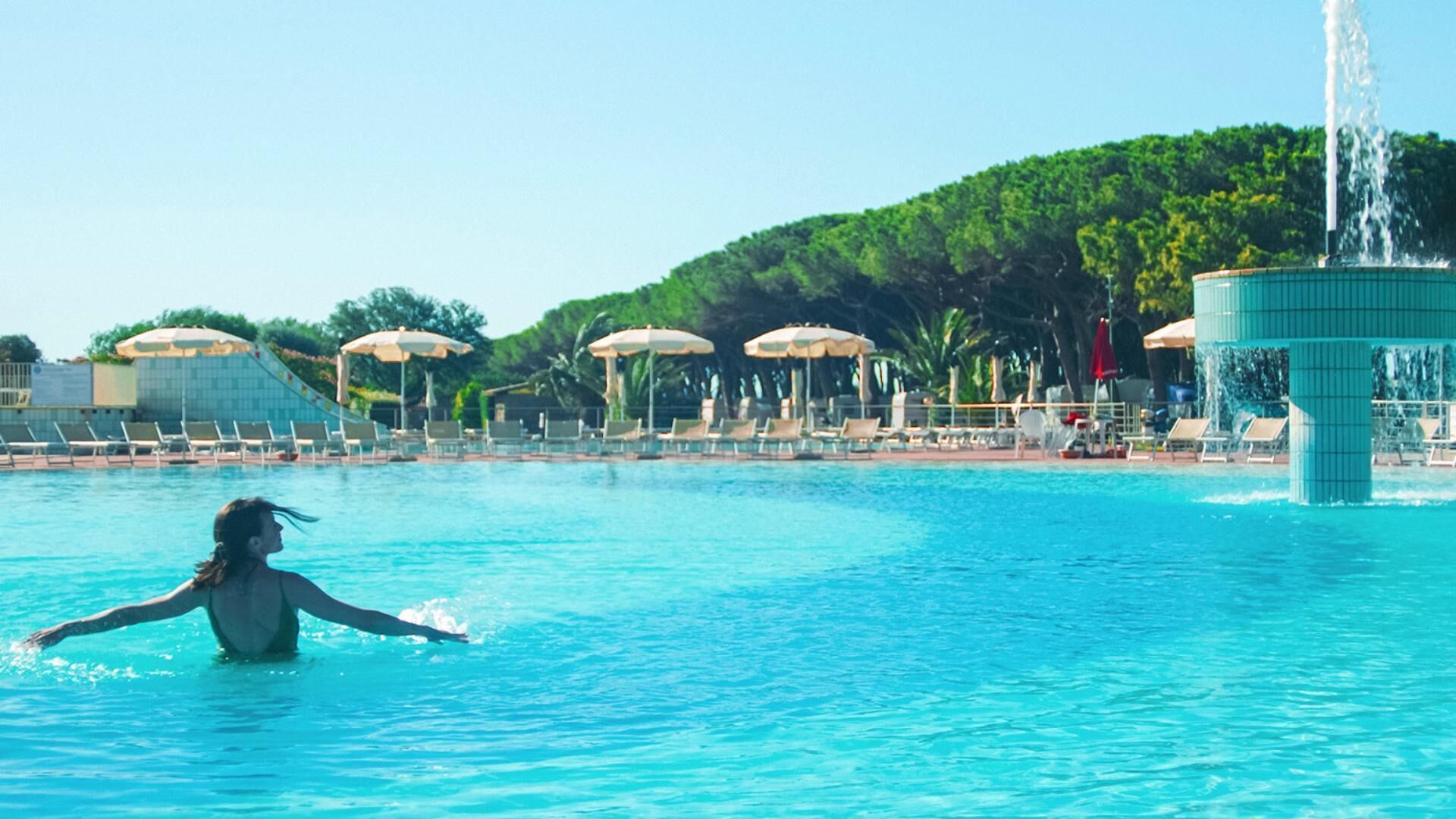 Une piscine avec des chaises longues et des parasols, une fontaine et une colline en arrière-plan.