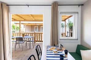 Ein gemütliches Zimmer mit einer Holzdachterrasse, einer Essecke mit Tisch und Stühlen, einem Fenster mit Vorhängen und einem grünen Sofa.