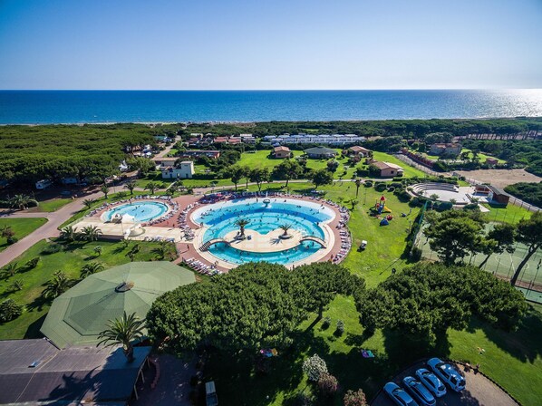 An aerial view of a resort with multiple swimming pools, a large grassy area, and a view of the sea.