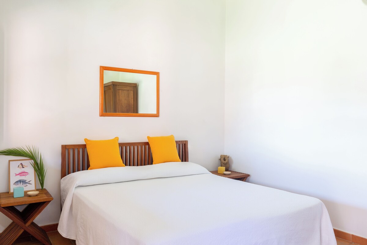 Un lit bien fait, avec une literie blanche et deux coussins orange vif, une tête de lit en bois, une petite table de chevet avec une lampe et, au mur, un tableau représentant des poissons.