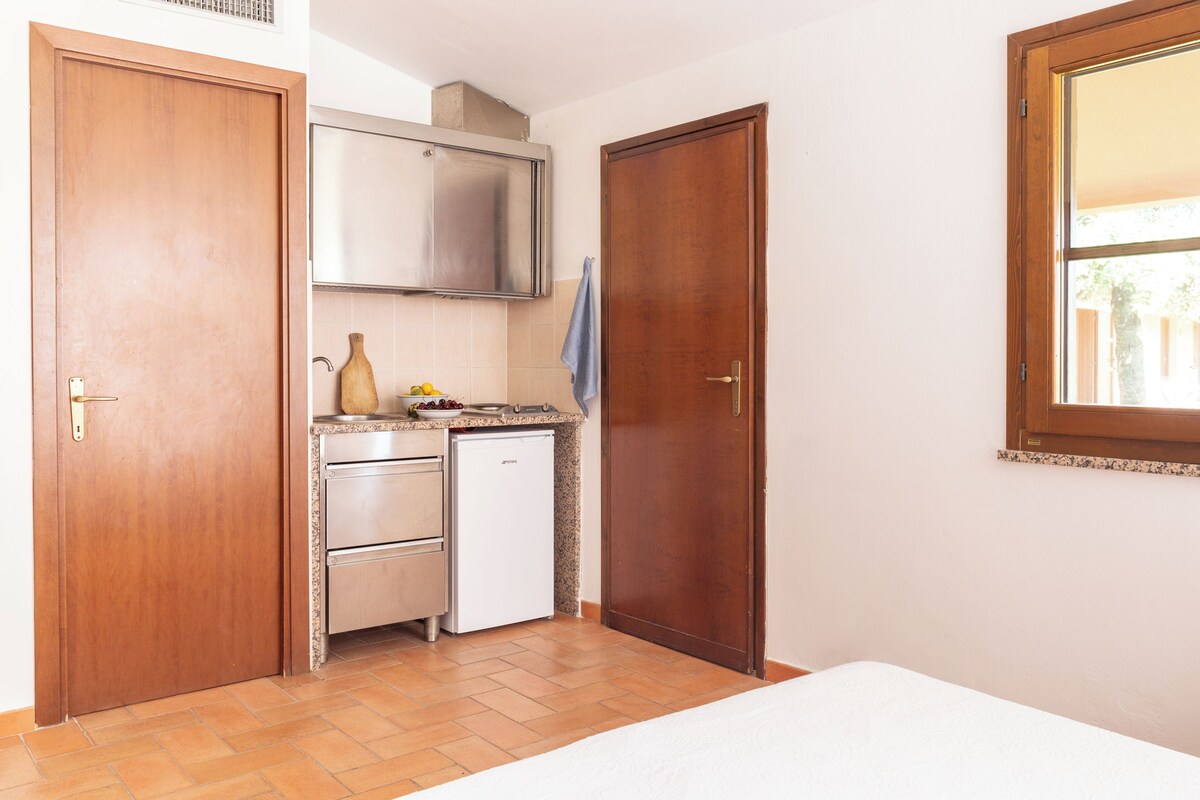 Eine kleine Küche mit einem Kühlschrank, einer Spüle und einem Herd. Links ist eine Holztür und rechts ein Fenster zu sehen.