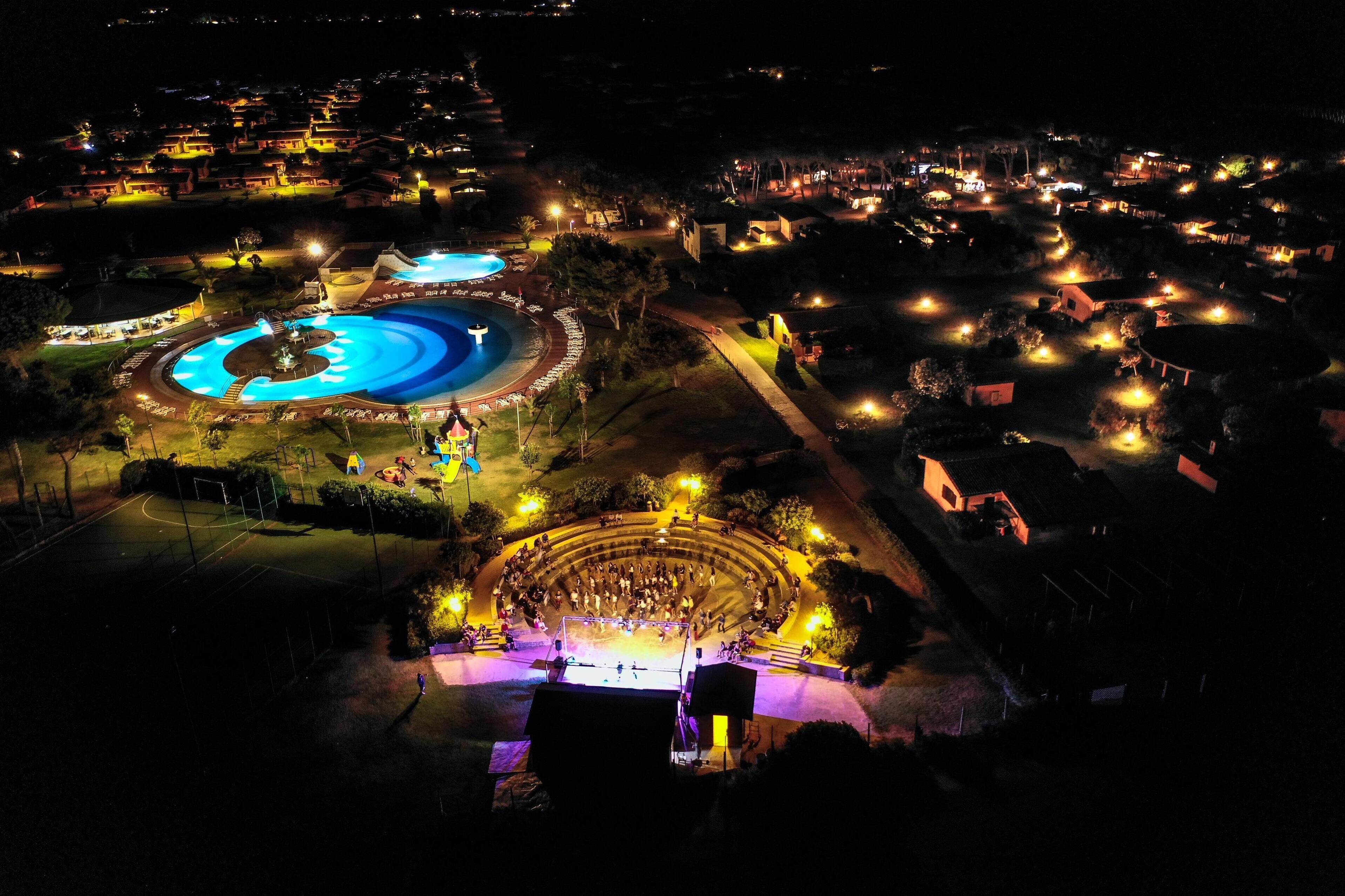 An aerial view of a resort with a large swimming pool, a circular amphitheater, and well-lit pathways.