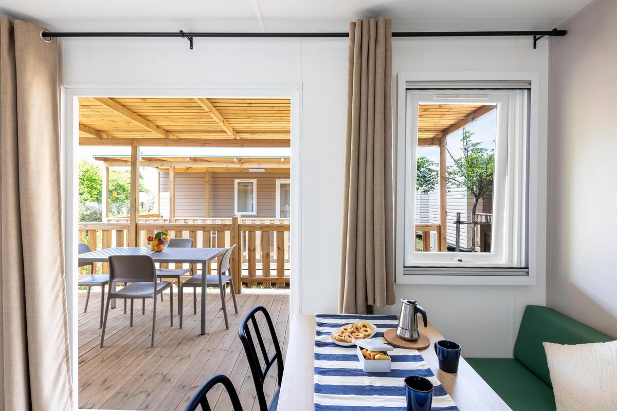 Une pièce chaleureuse avec une terrasse en bois, une table à manger avec des chaises, une fenêtre avec des rideaux et un canapé vert.