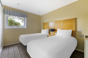 A hotel room with two beds, a window with a plaid valance, a wooden headboard, a bedside lamp, and a radiator.