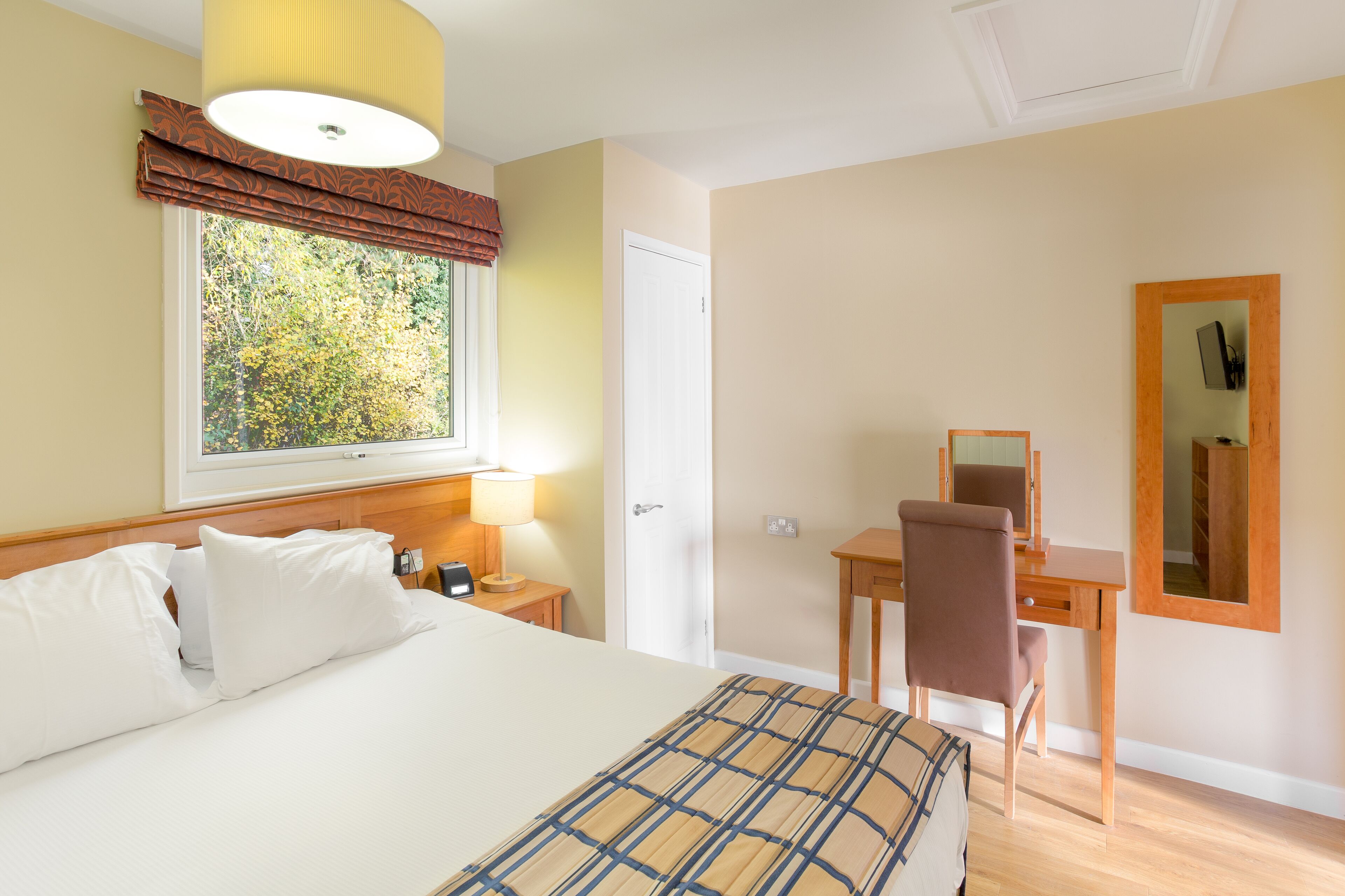 A hotel room with a bed, a desk, a chair, a lamp, a window with a view of trees, and a mirror on the wall.