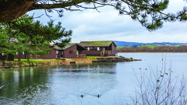 A cabin-style building by a lake with a dock, surrounded by trees and hills in the background. - Pine Lake Resort (Carnforth)