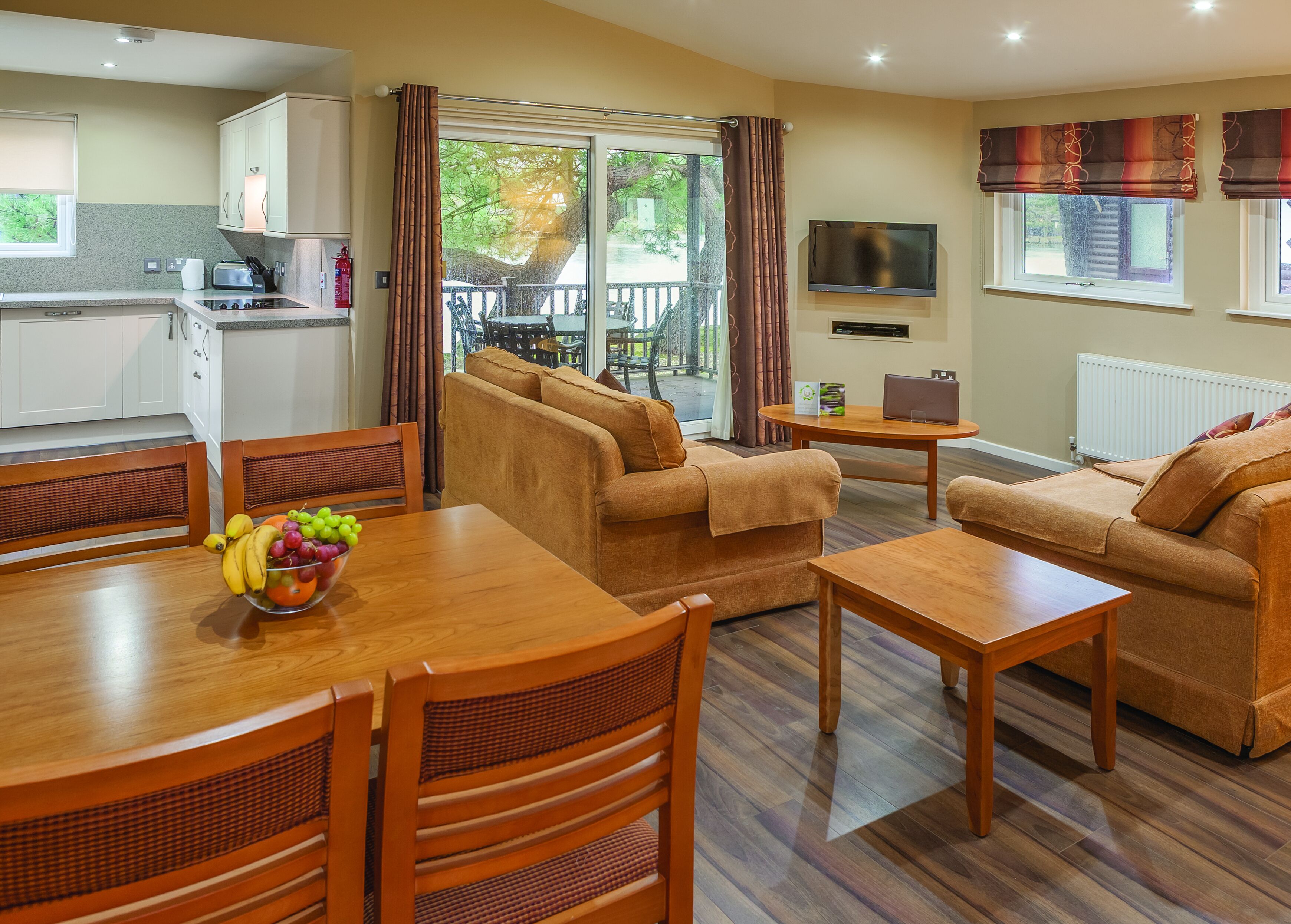 A modern living room with a sofa set, a dining table with fruit, and a kitchen area with cabinets and a TV.
