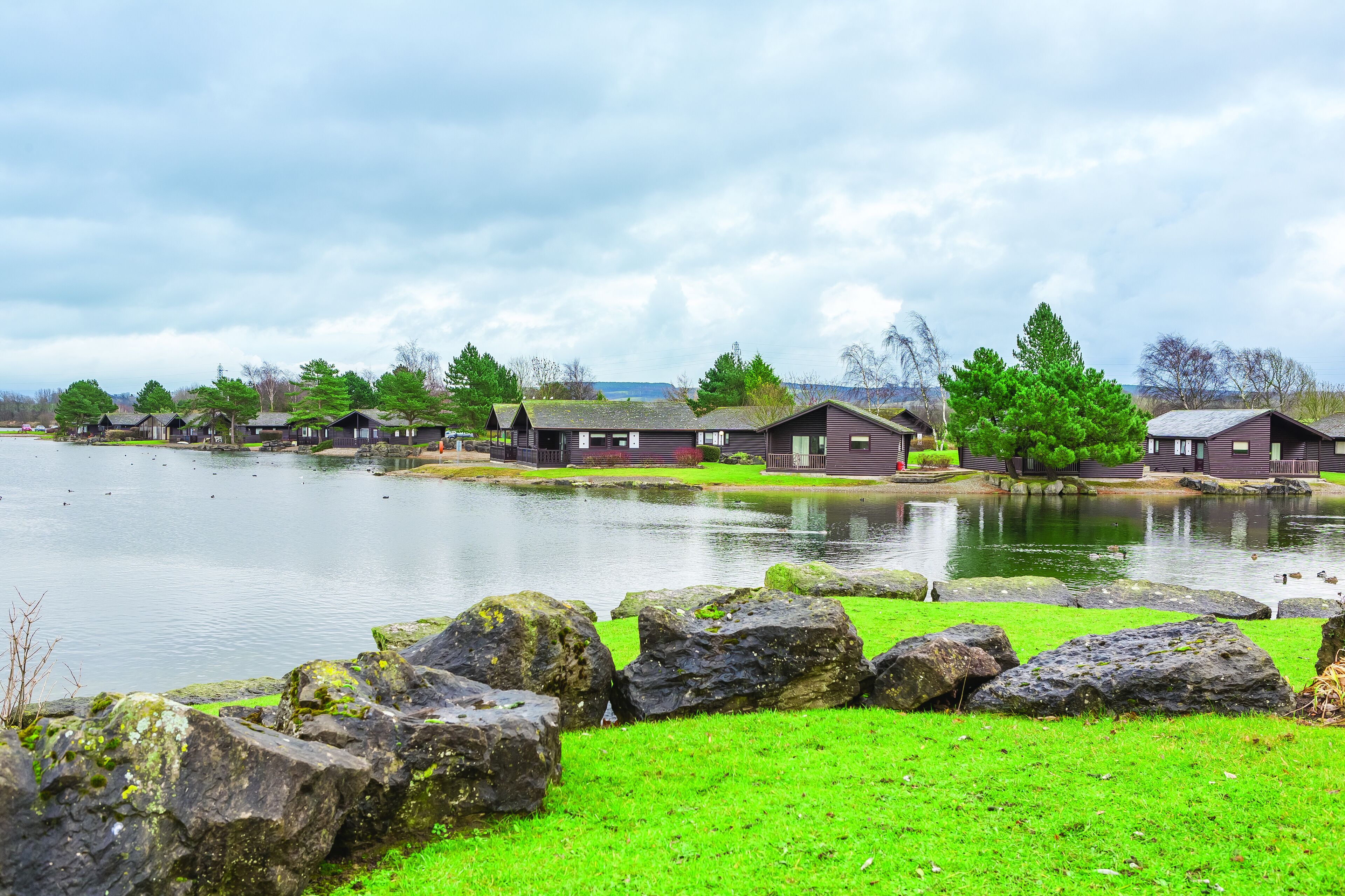 A row of cabins by a lake with a grassy foreground and large rocks.