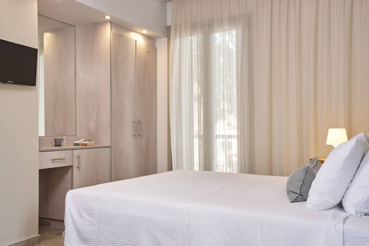A hotel room with a bed, a TV mounted on the wall, a desk with a lamp, and a window with curtains.