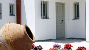 Un edifici blanc amb una porta i dues finestres, una olla de fang gran, i flors de colors al primer pla.