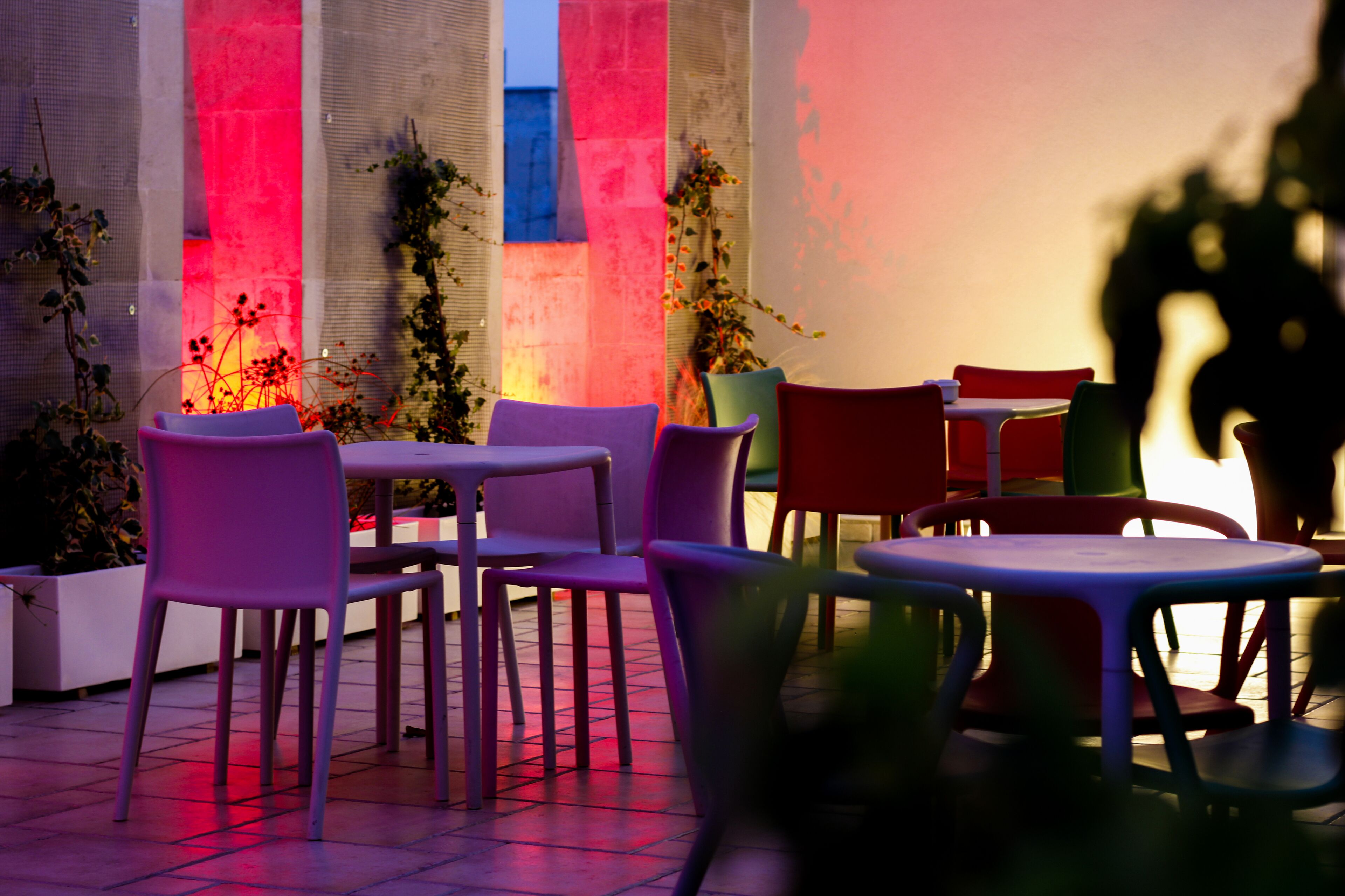 An outdoor seating area with tables and chairs, surrounded by plants and a textured wall.